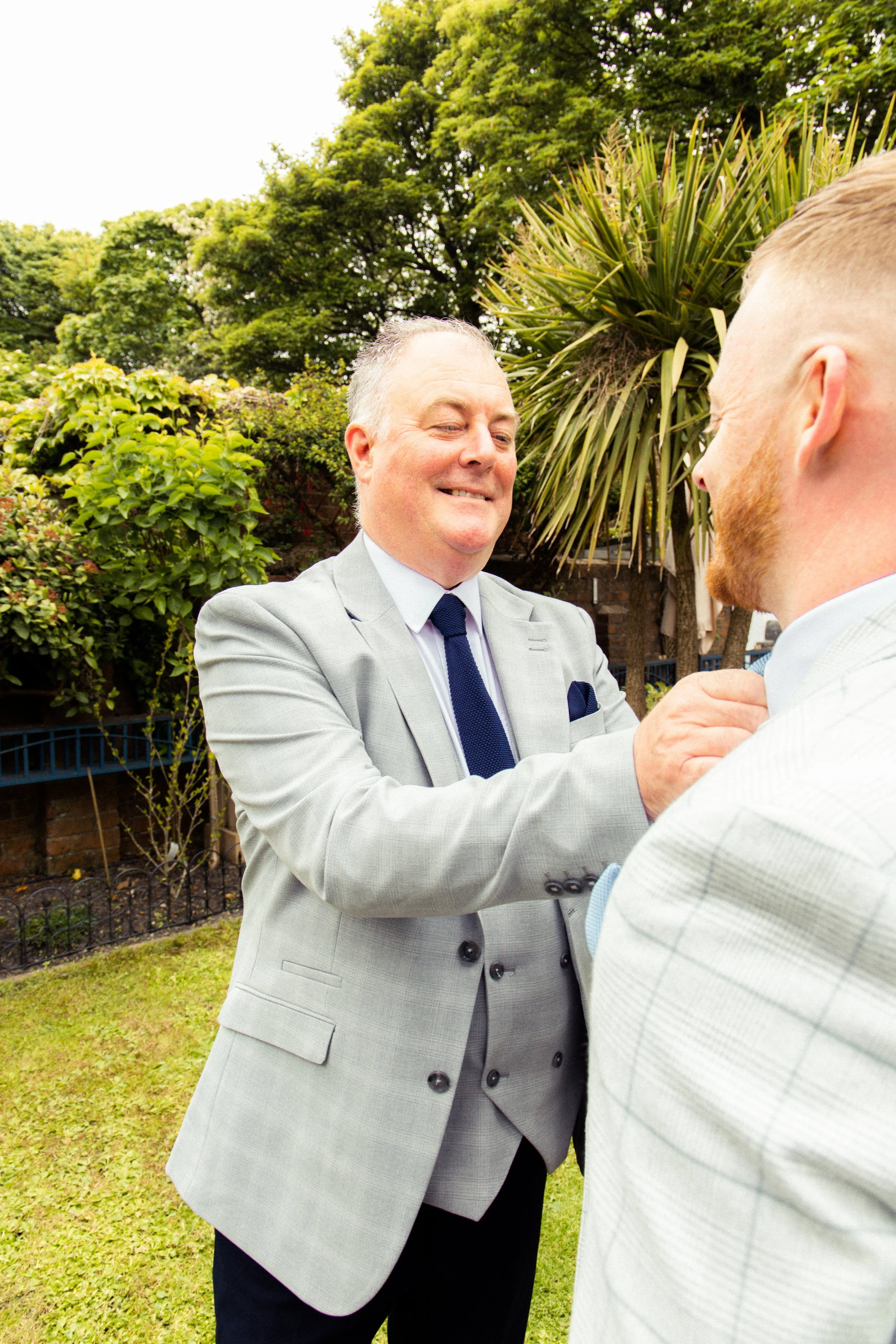 A man in a light gray suit and blue tie adjusting another man’s tie outdoors, with green trees and bushes in the background.
