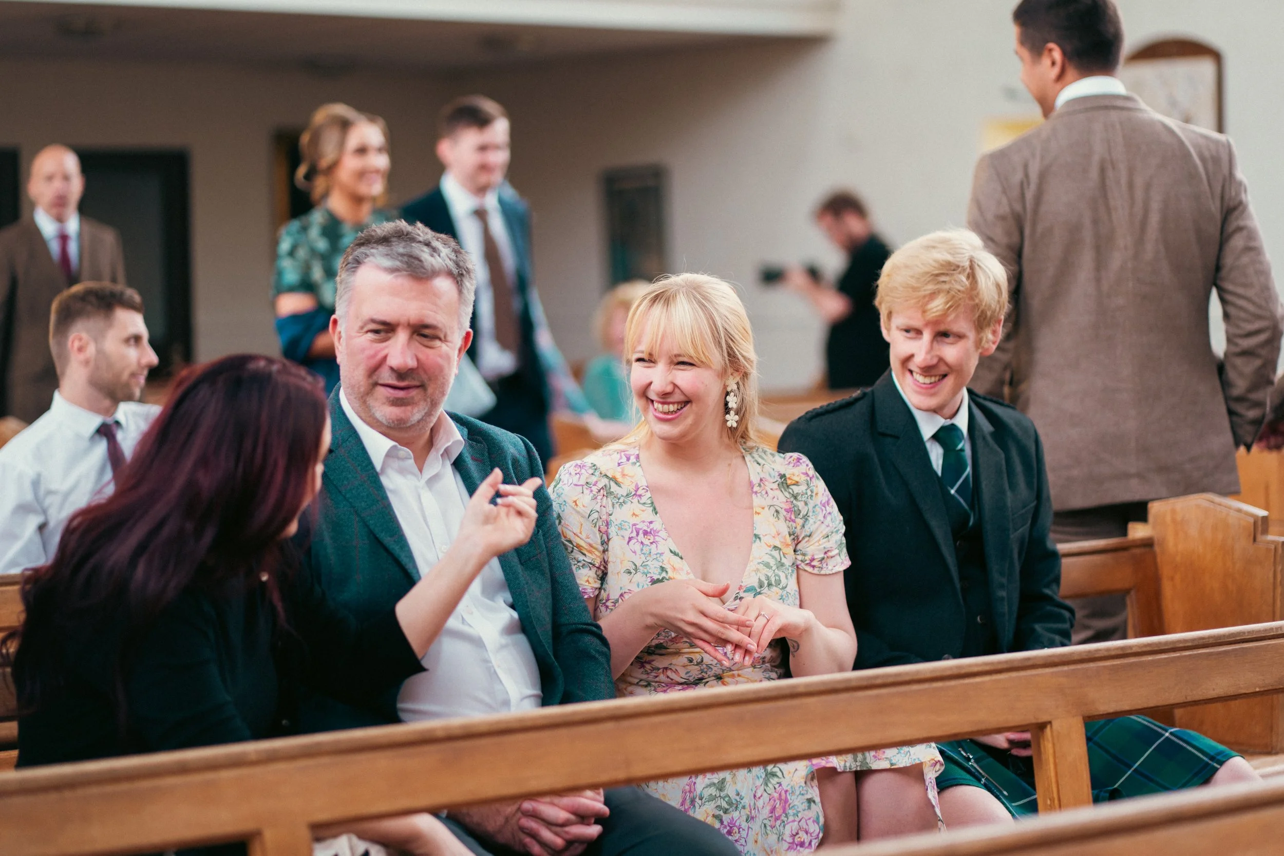 People sitting on pews in a church or event hall, smiling and engaging in conversation, with others standing in the background.