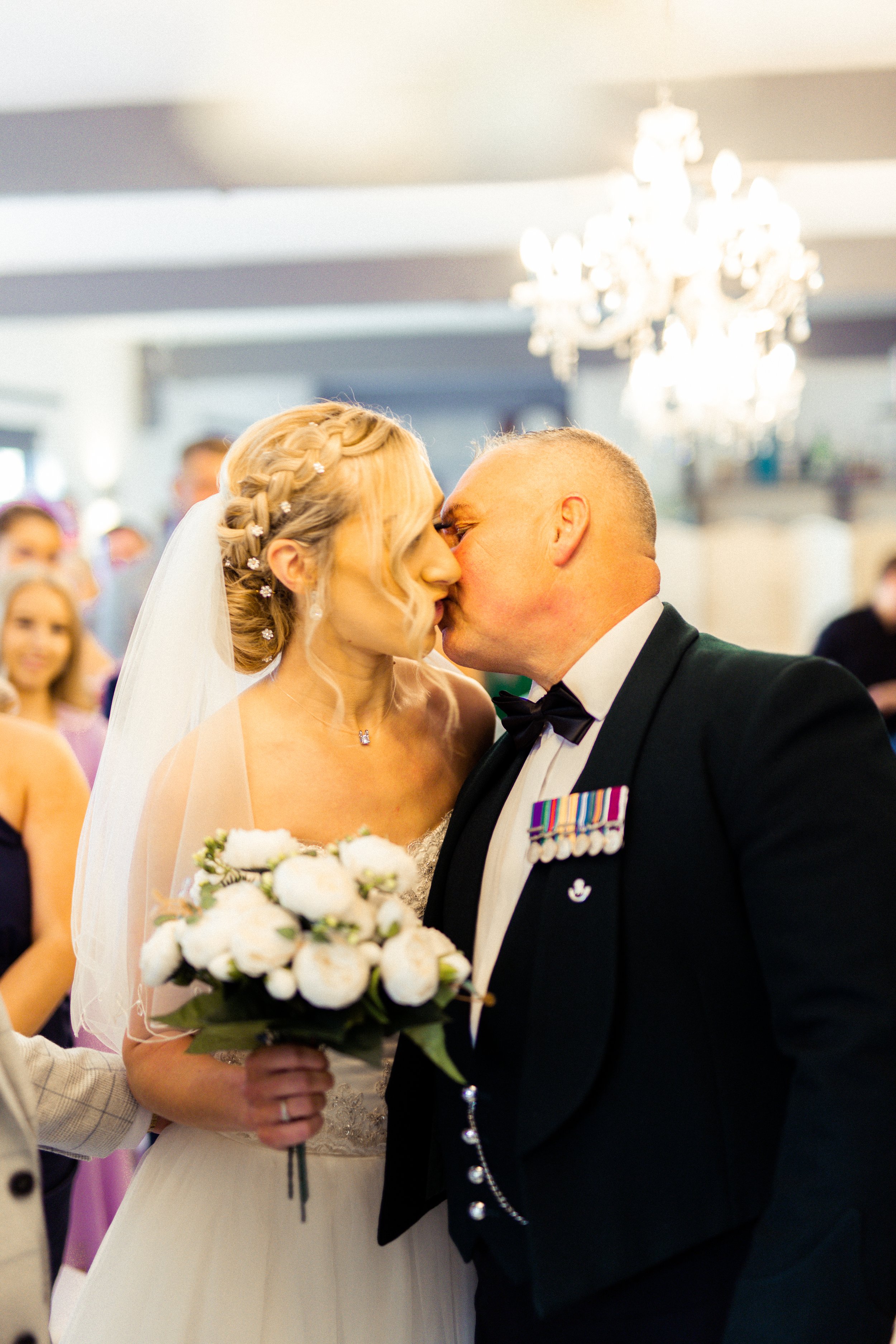 A bride and a groom share a kiss during their wedding ceremony, with the bride holding a bouquet of white flowers and wearing a white wedding gown and veil, while the groom is in a black tuxedo with medals on his chest.