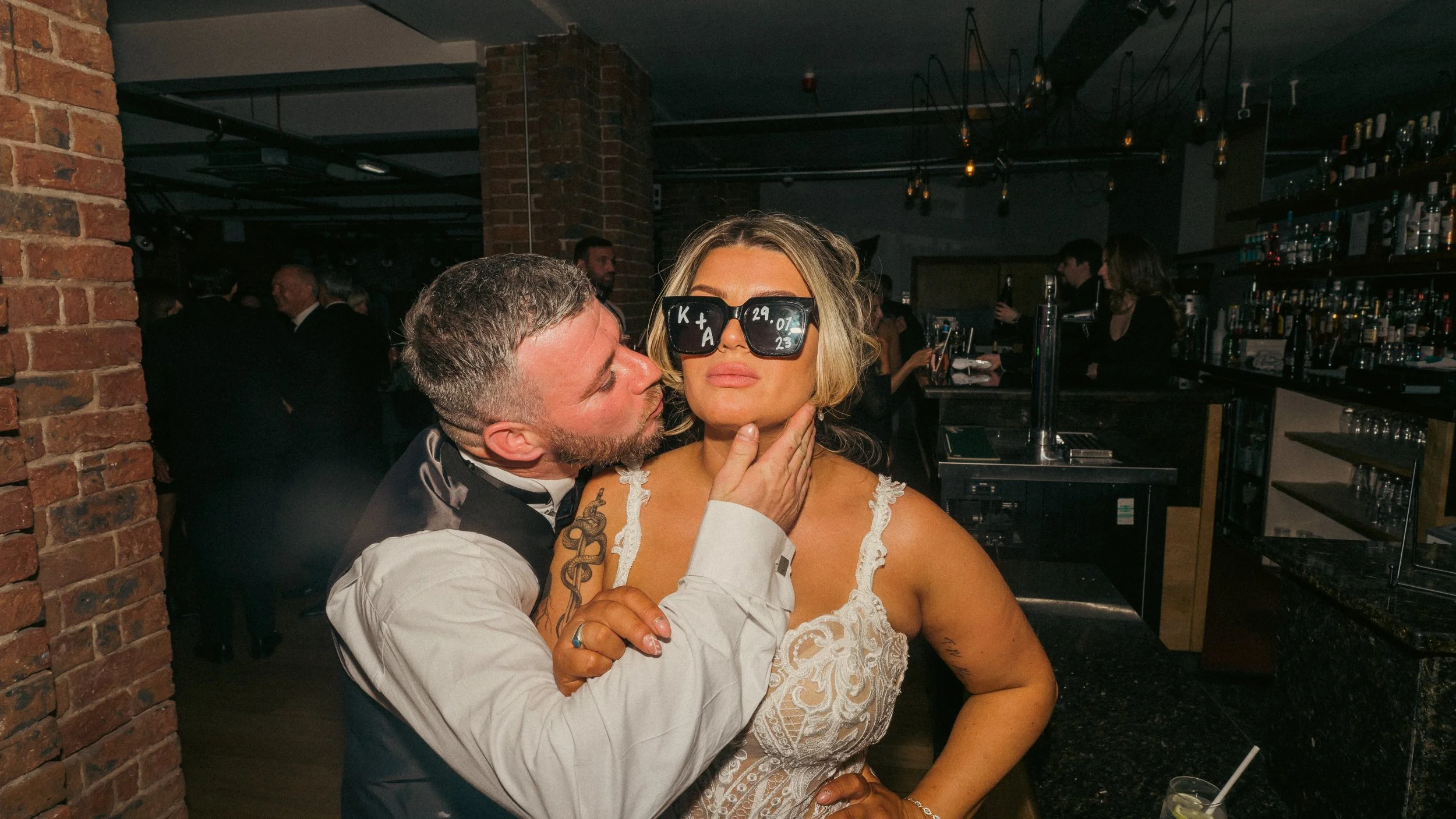 A man kisses a woman on the cheek at a bar, with the woman wearing large sunglasses and a lace dress, while the man is in a tuxedo, with a bar scene in the background.