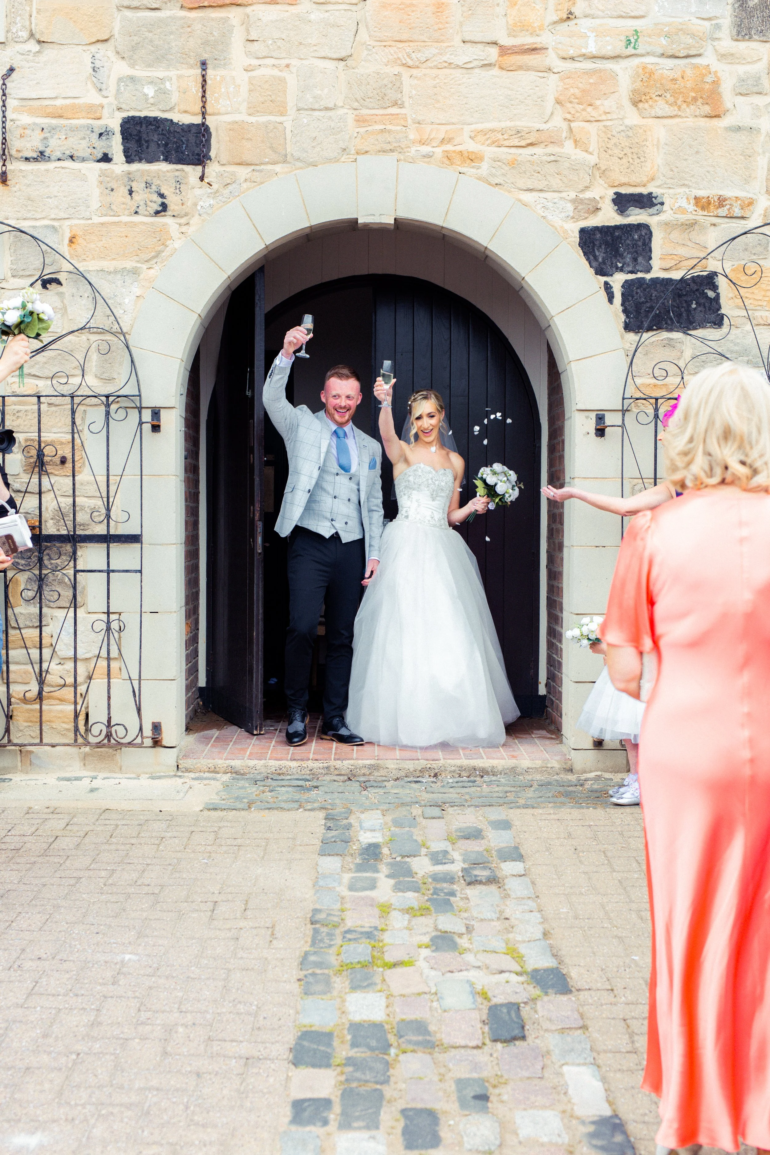 A newlywed couple celebrating as they exit a stone building, holding glasses of champagne, while guests throw flower petals.
