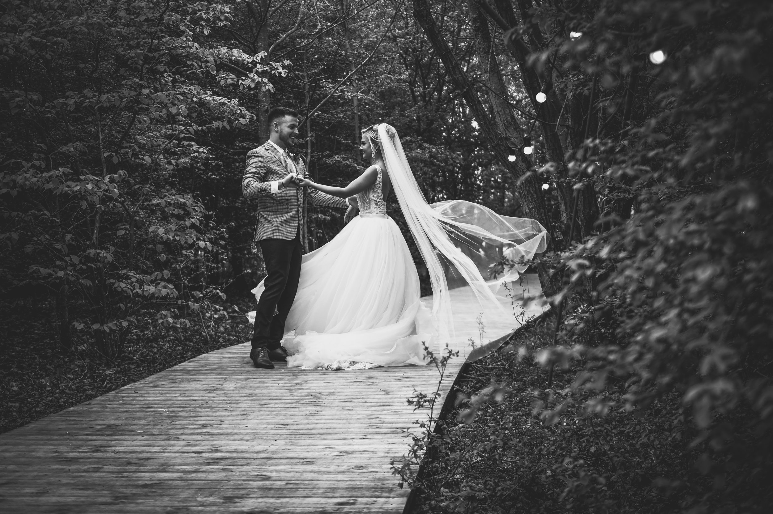 A black and white wedding photo of a couple dancing on a wooden pathway in a forested area, with the bride in a flowing gown and veil, and the groom in a checked suit jacket. The bride's veil is caught in the wind, and they are smiling at each other.