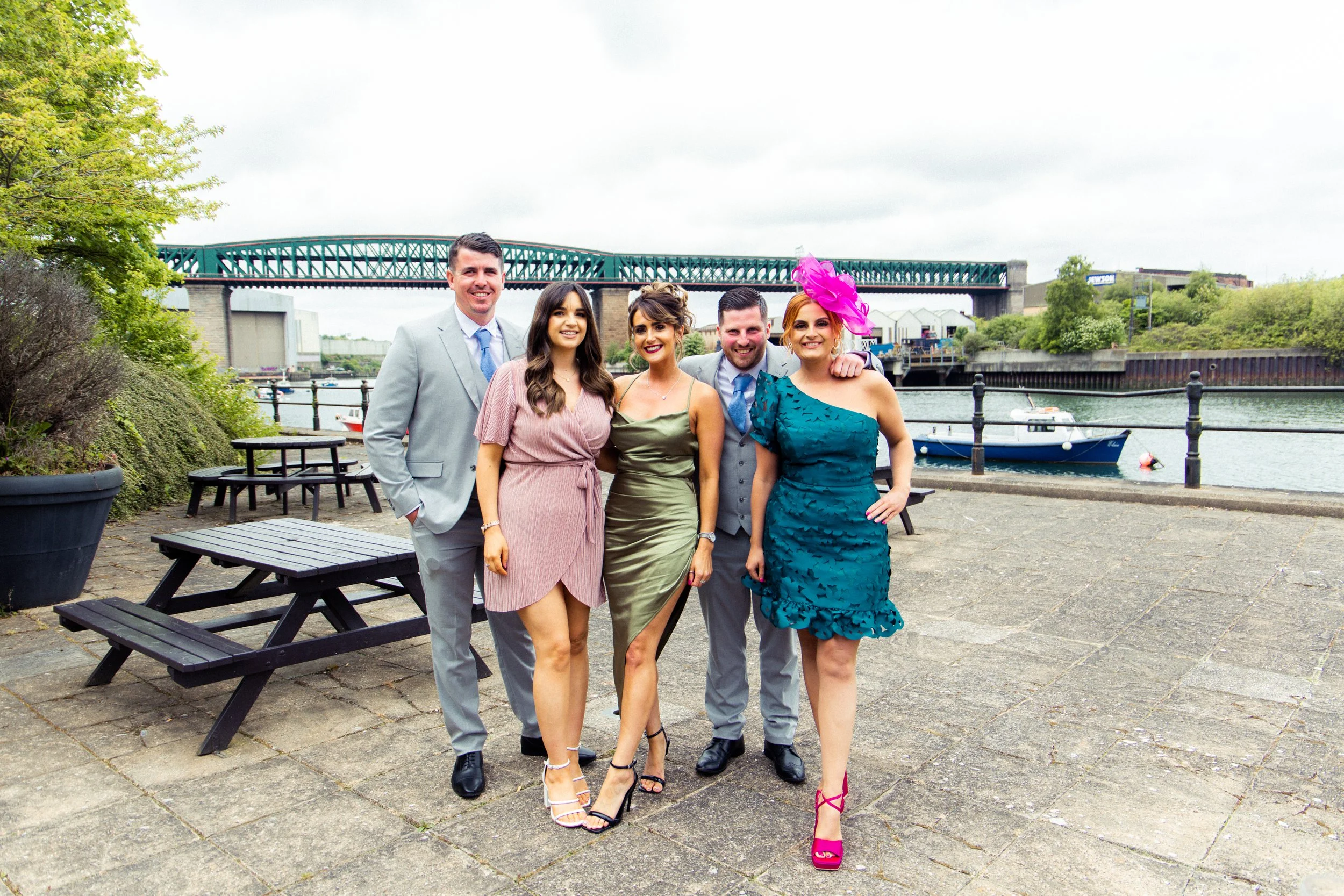 Five people standing outdoors near water with boats and a bridge in the background, dressed in formal attire