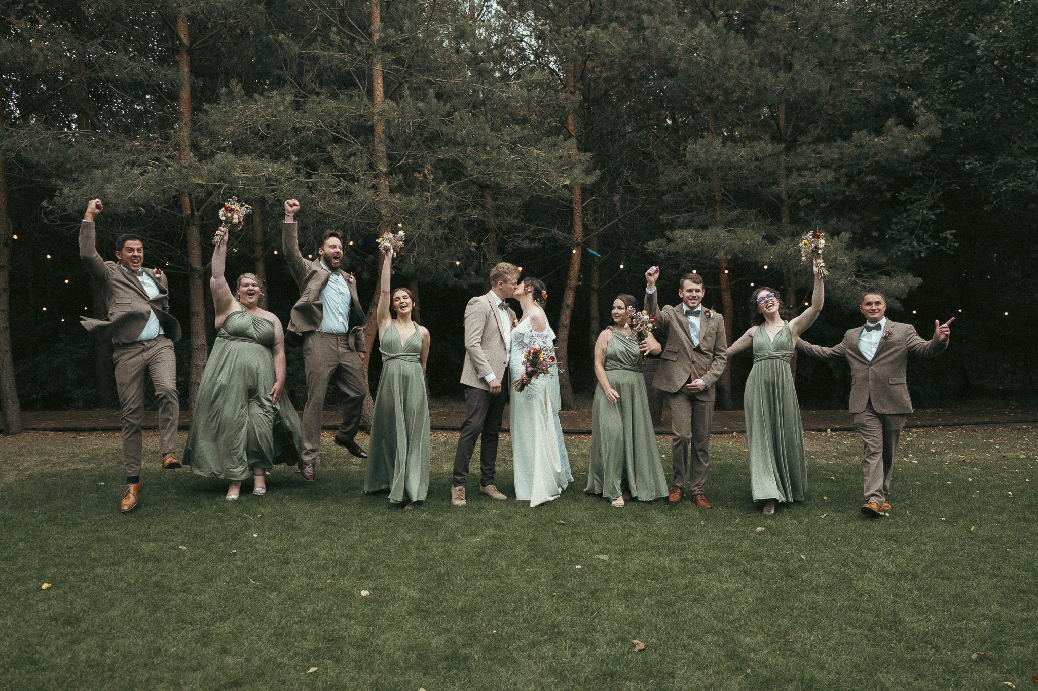 A wedding party standing outdoors on grass in front of trees, including the bride and groom kissing, with bridesmaids in green dresses and groomsmen in brown suits celebrating and holding flowers.