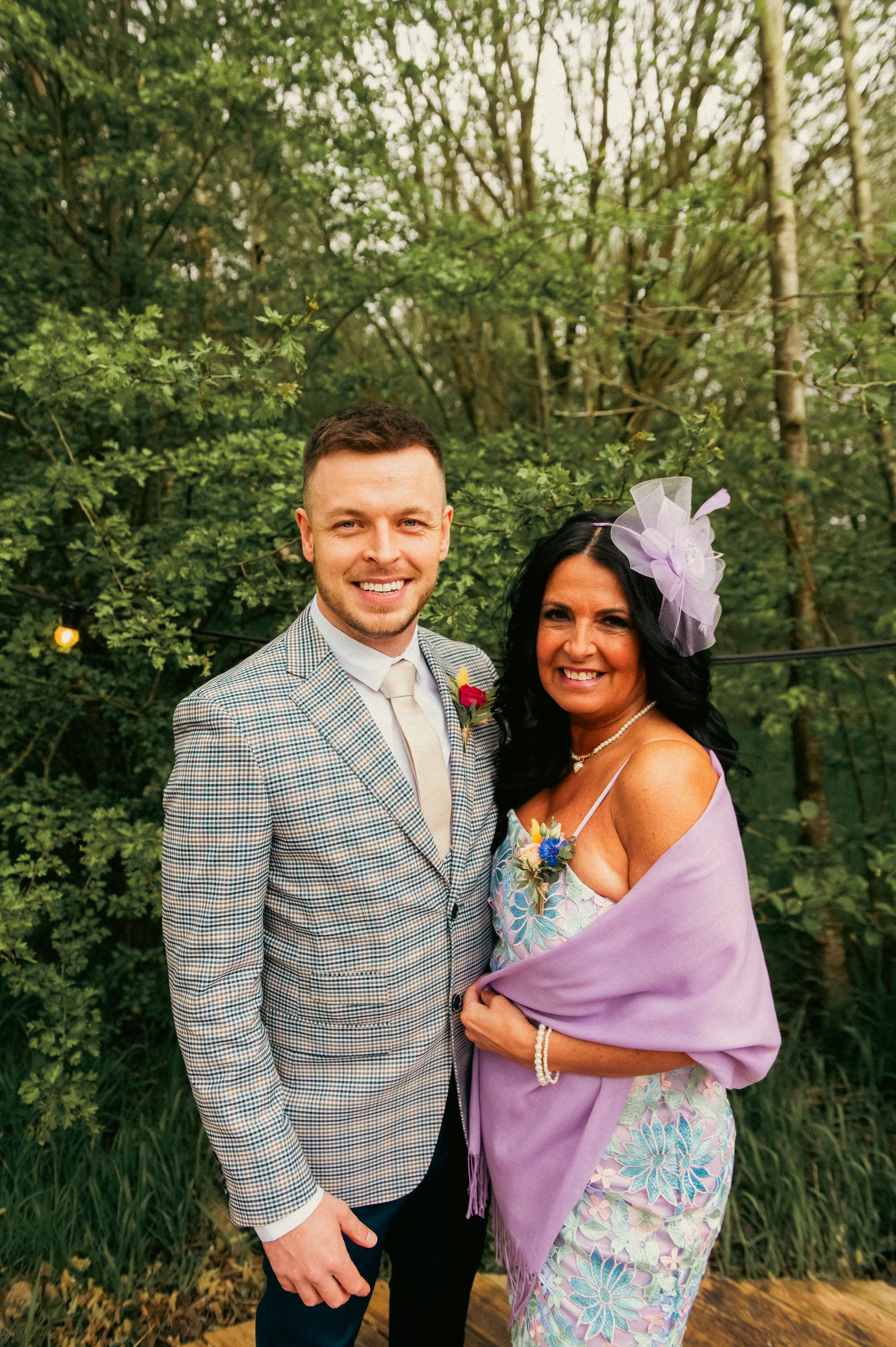 A man and a woman smiling and embracing outdoors among green trees. The man is wearing a checkered suit and tie, and the woman is in a floral dress with a lavender shawl, a pearl bracelet, and a purple fascinator hat.
