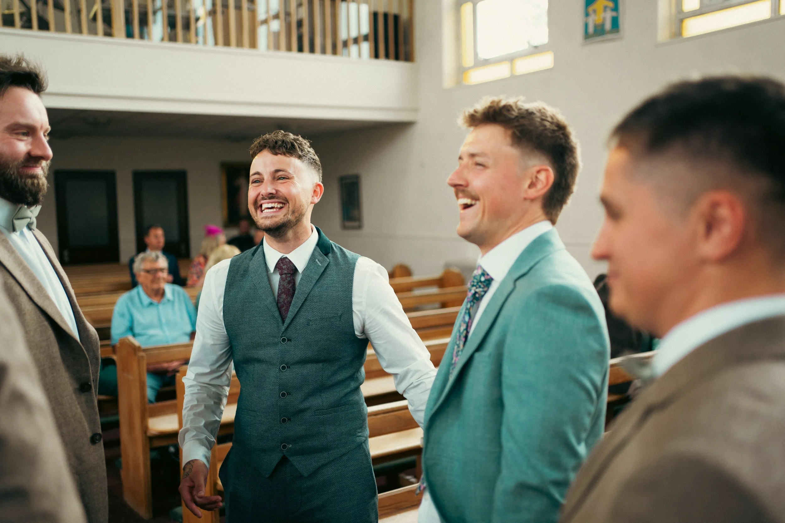 Four men dressed in suits having a conversation and smiling inside a church during a wedding ceremony, with pews and guests in the background.