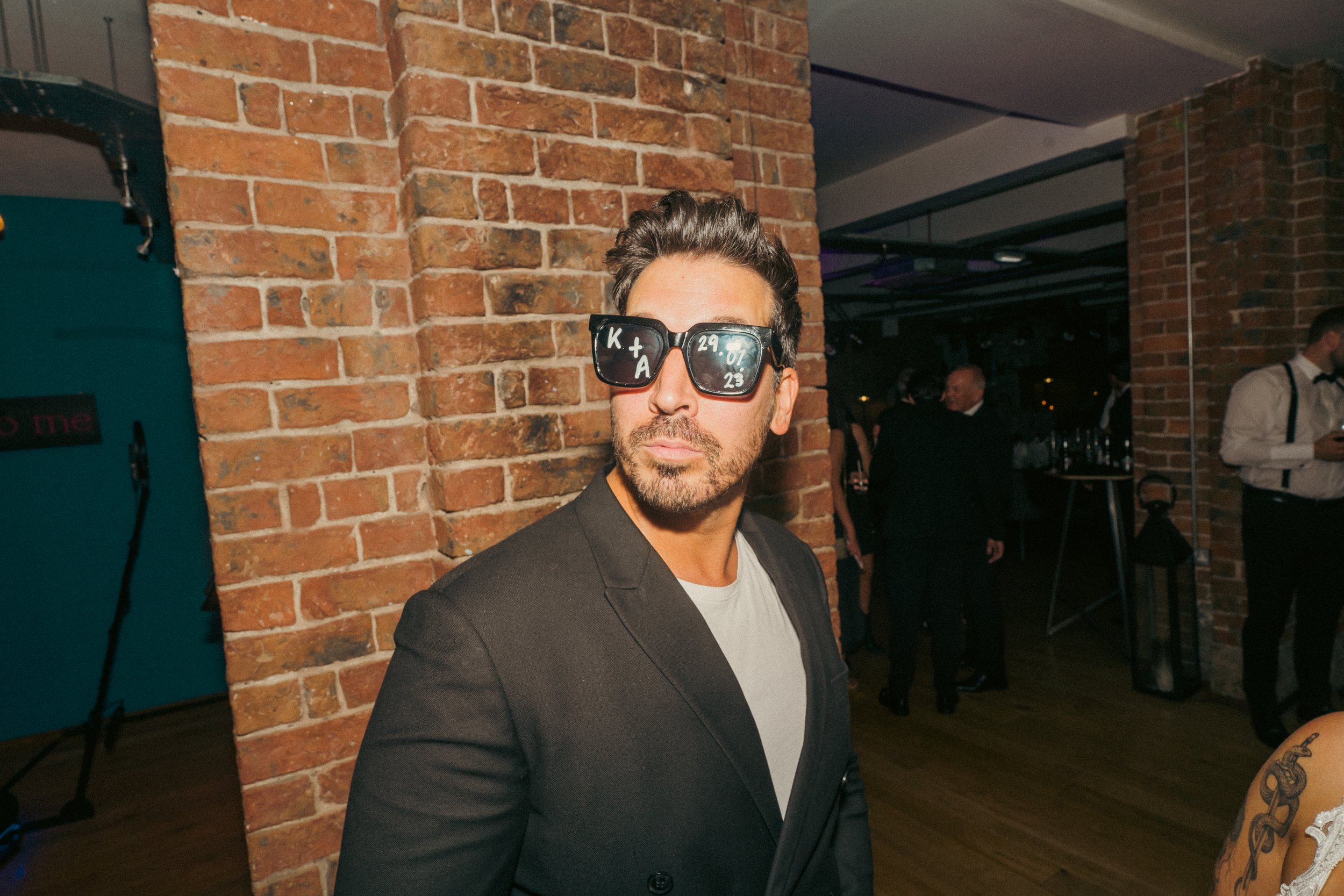 A man with dark hair and beard wearing large sunglasses, a black jacket, and a light-colored shirt, standing indoors with an exposed brick wall behind him at a social event.