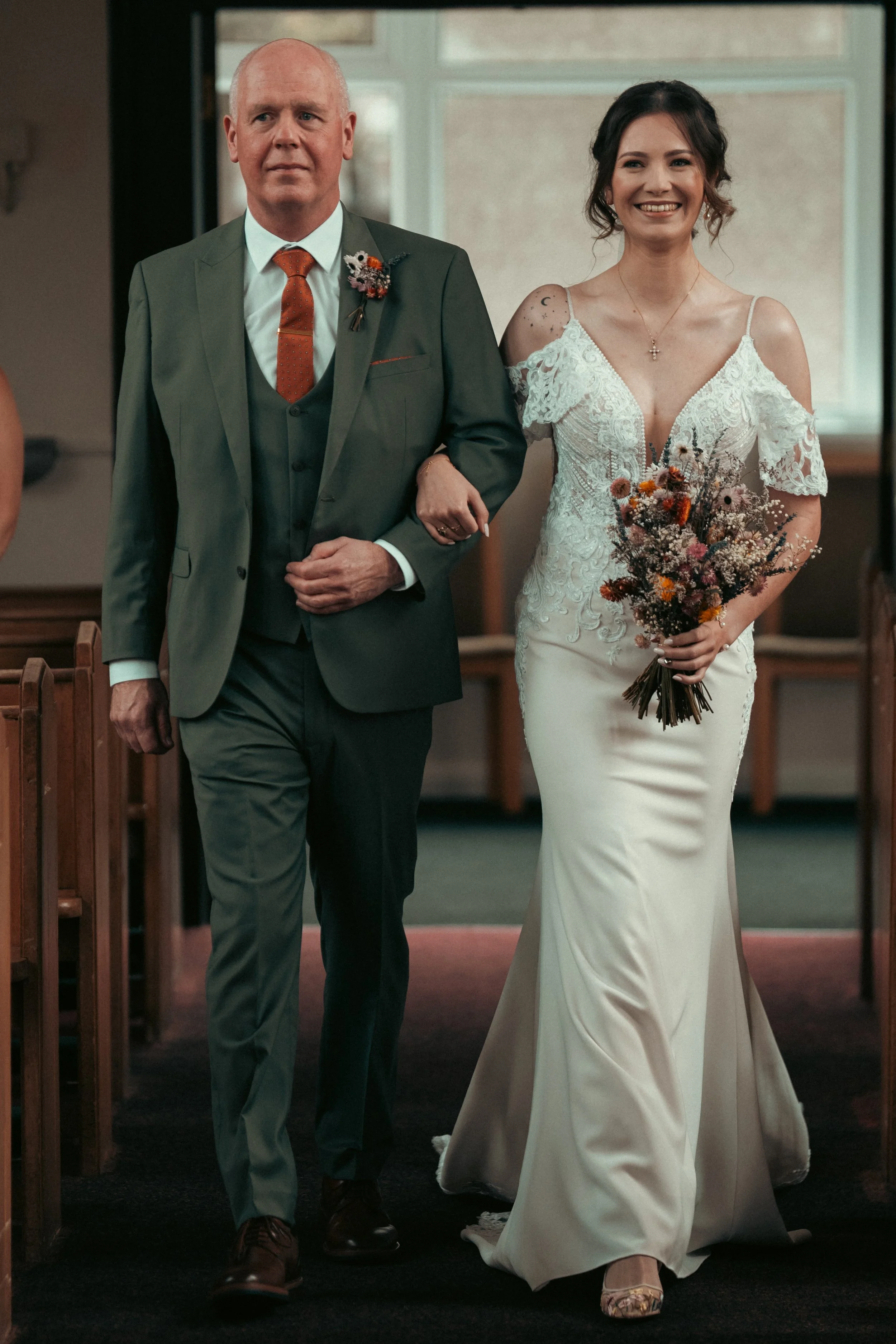 A bride in a white wedding dress walking down the aisle with an older man, likely her father, during a wedding ceremony inside a church.