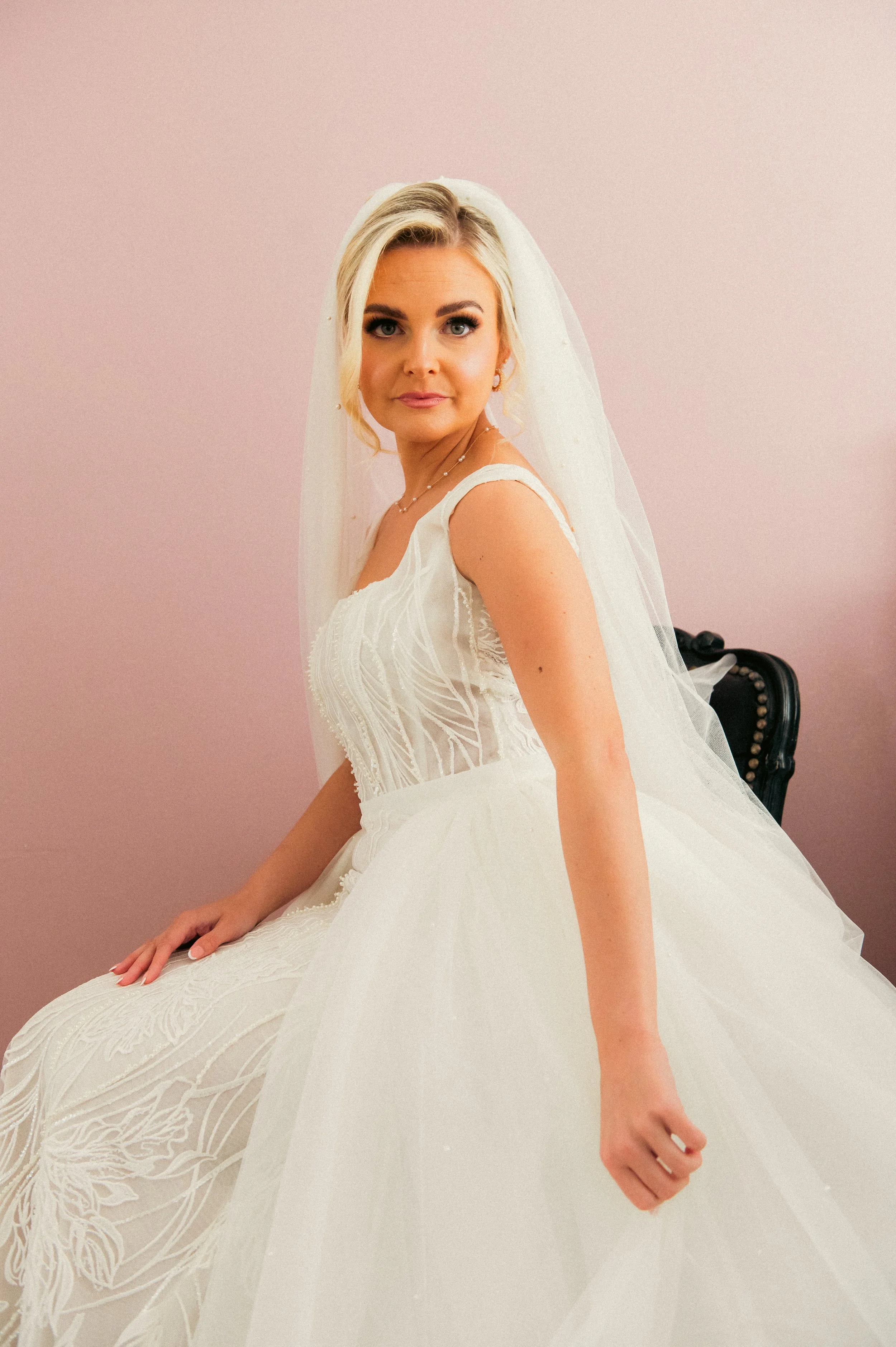 A woman in a white wedding dress with lace details, sitting on a chair, with a pink wall behind her and a sheer veil over her blonde hair.