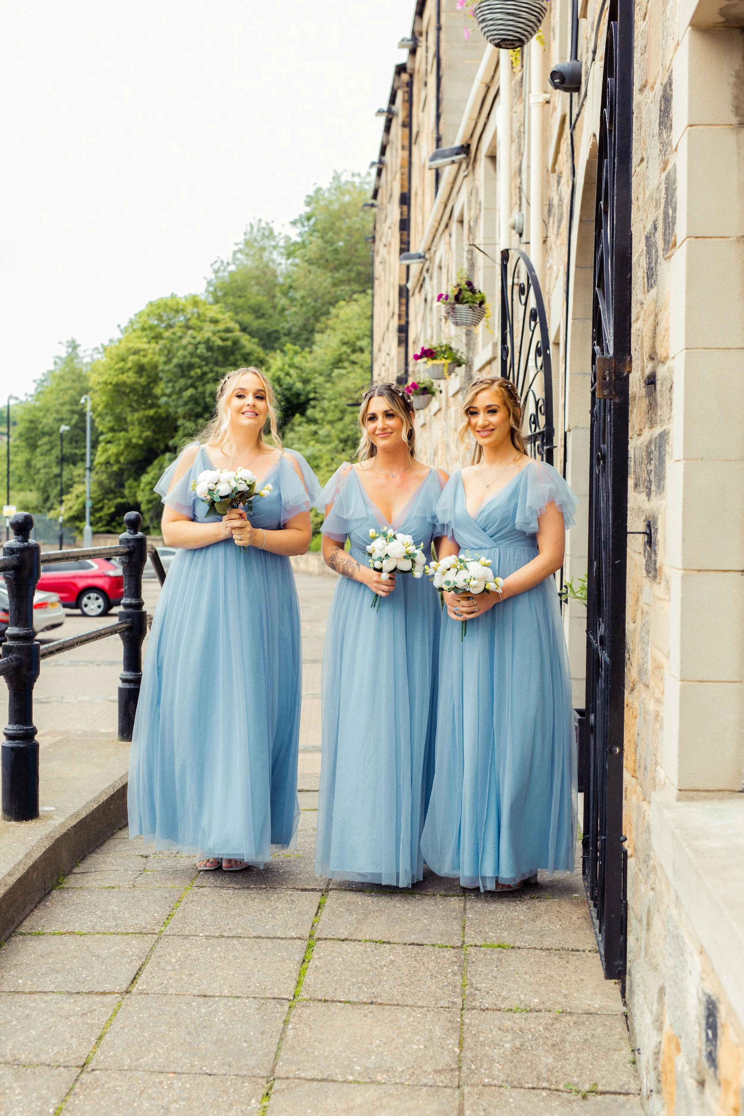 Three women in matching light blue dresses holding white floral bouquets standing on a sidewalk near a stone building with potted flowers.