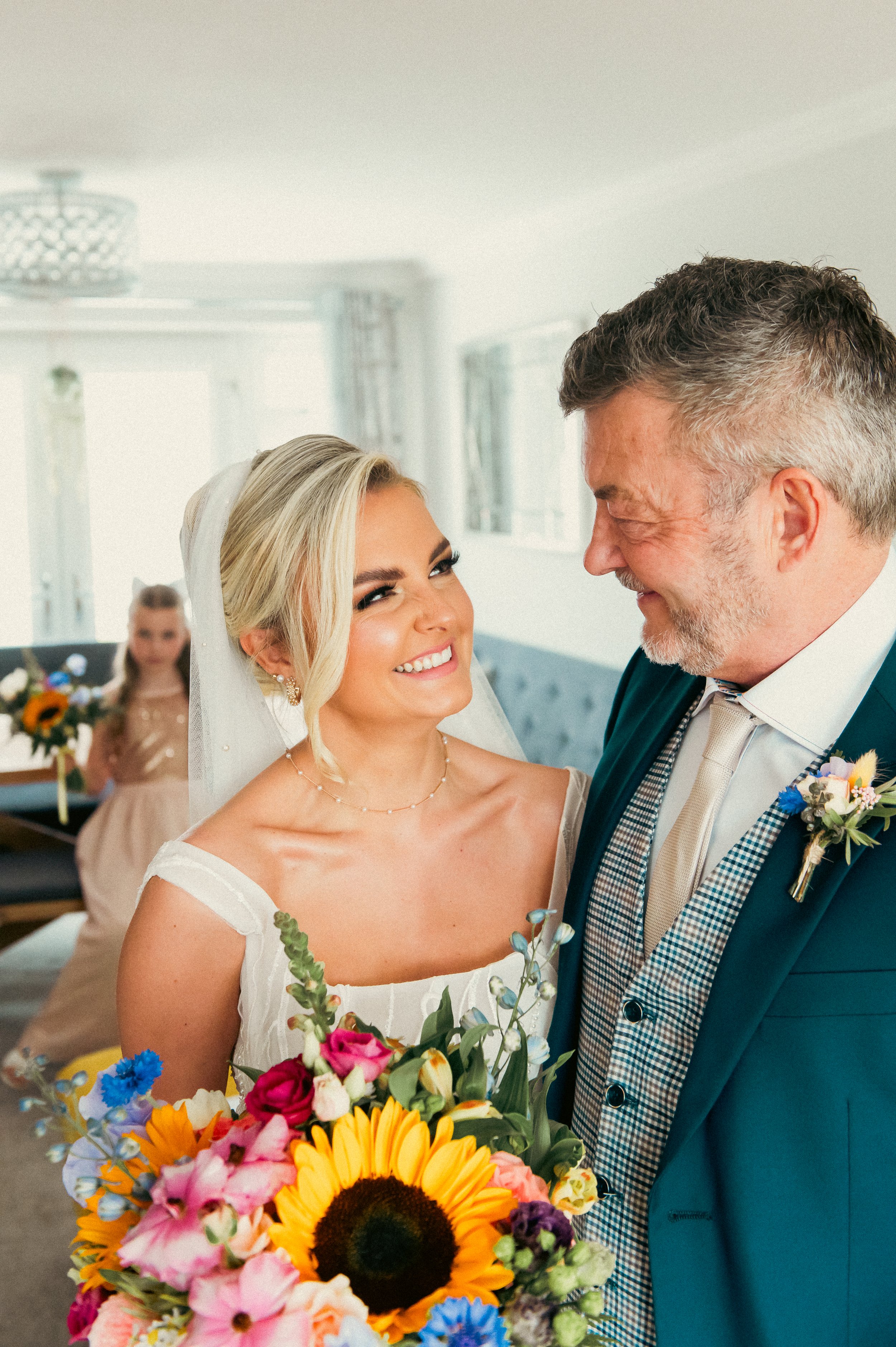A bride smiling at a man, holding a bouquet of colorful flowers in a bright room with a woman in the background.