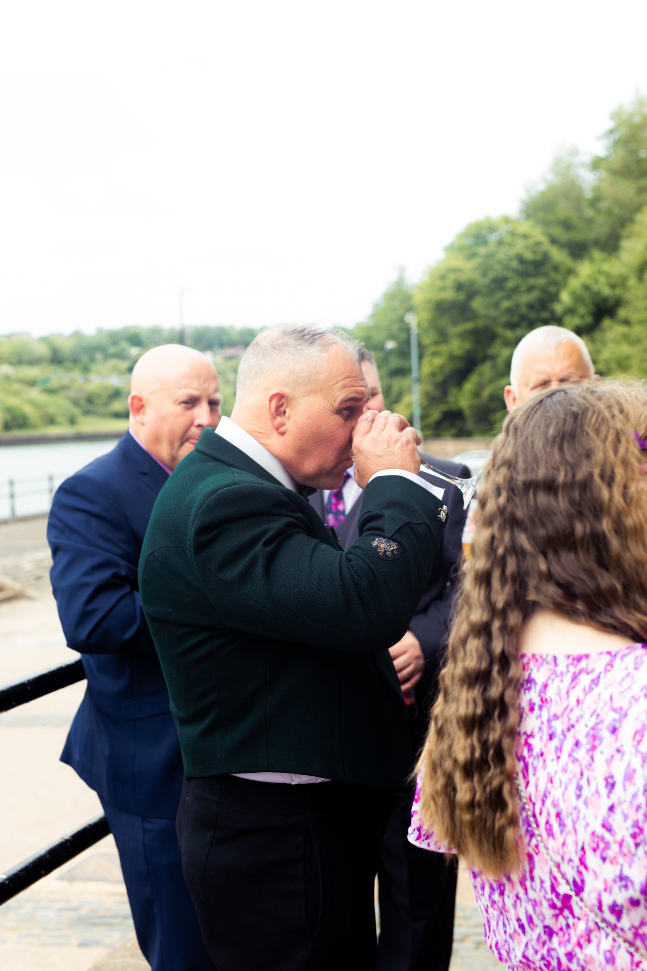 Man in black suit taking a sip from a glass during an outdoor event, with others around him near a waterfront and green trees in the background.