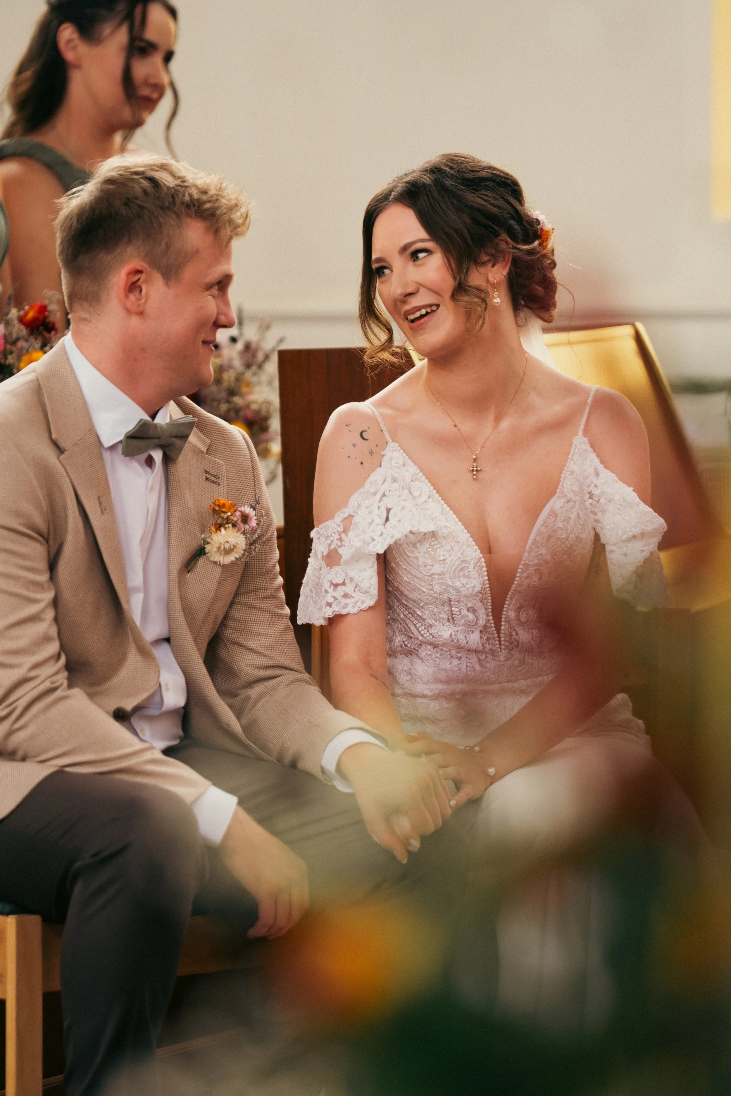 A woman in a wedding dress holding hands with a man in a beige suit during a wedding ceremony, with another woman in the background.