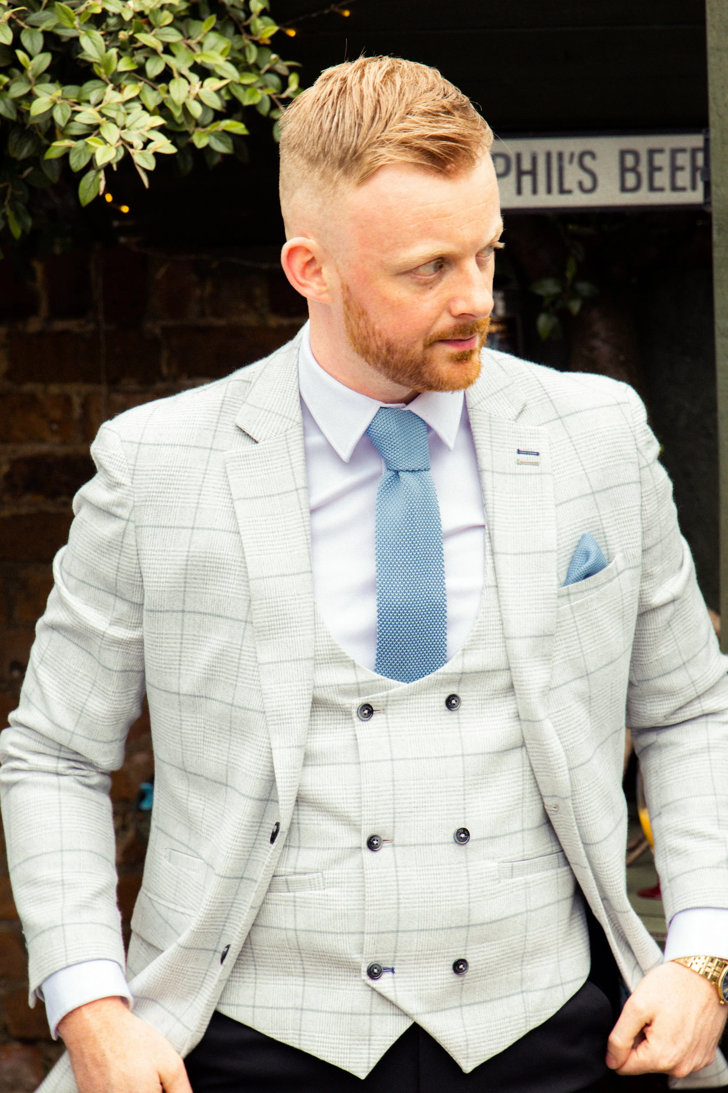 A man with red hair and a beard wearing a light gray plaid suit with a white shirt, light blue tie, and light blue pocket square. There is a sign in the background that says "PHIL'S BEER" and some greenery.