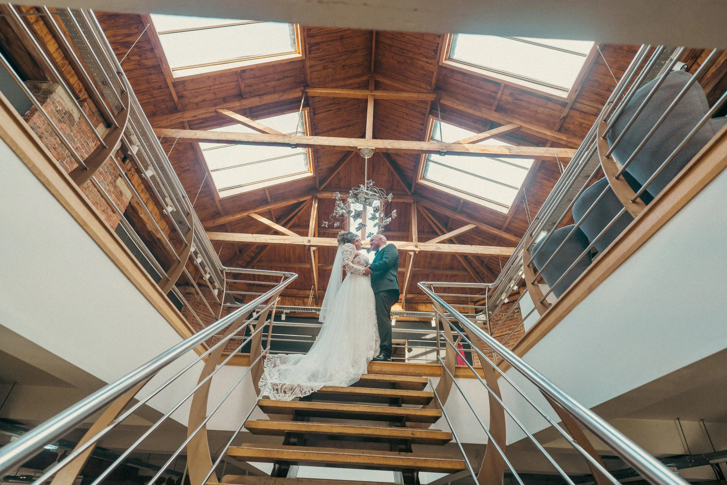 A bride and groom standing on a wooden staircase inside a building with a high, wooden gabled ceiling with large skylights. They are holding hands and facing each other in a romantic moment.
