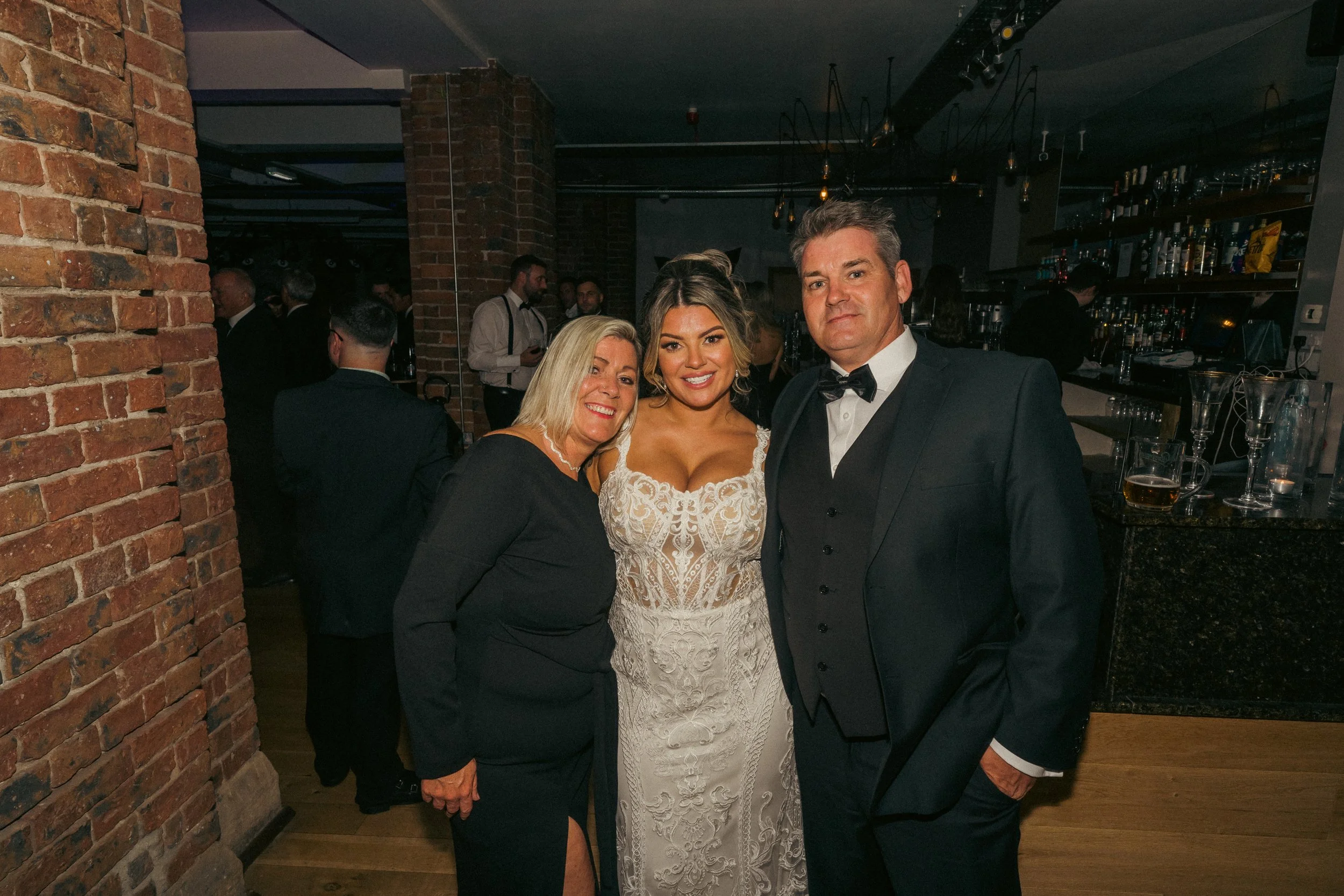Three people dressed in formal attire at a social event, standing inside a venue with brick walls and a bar in the background.