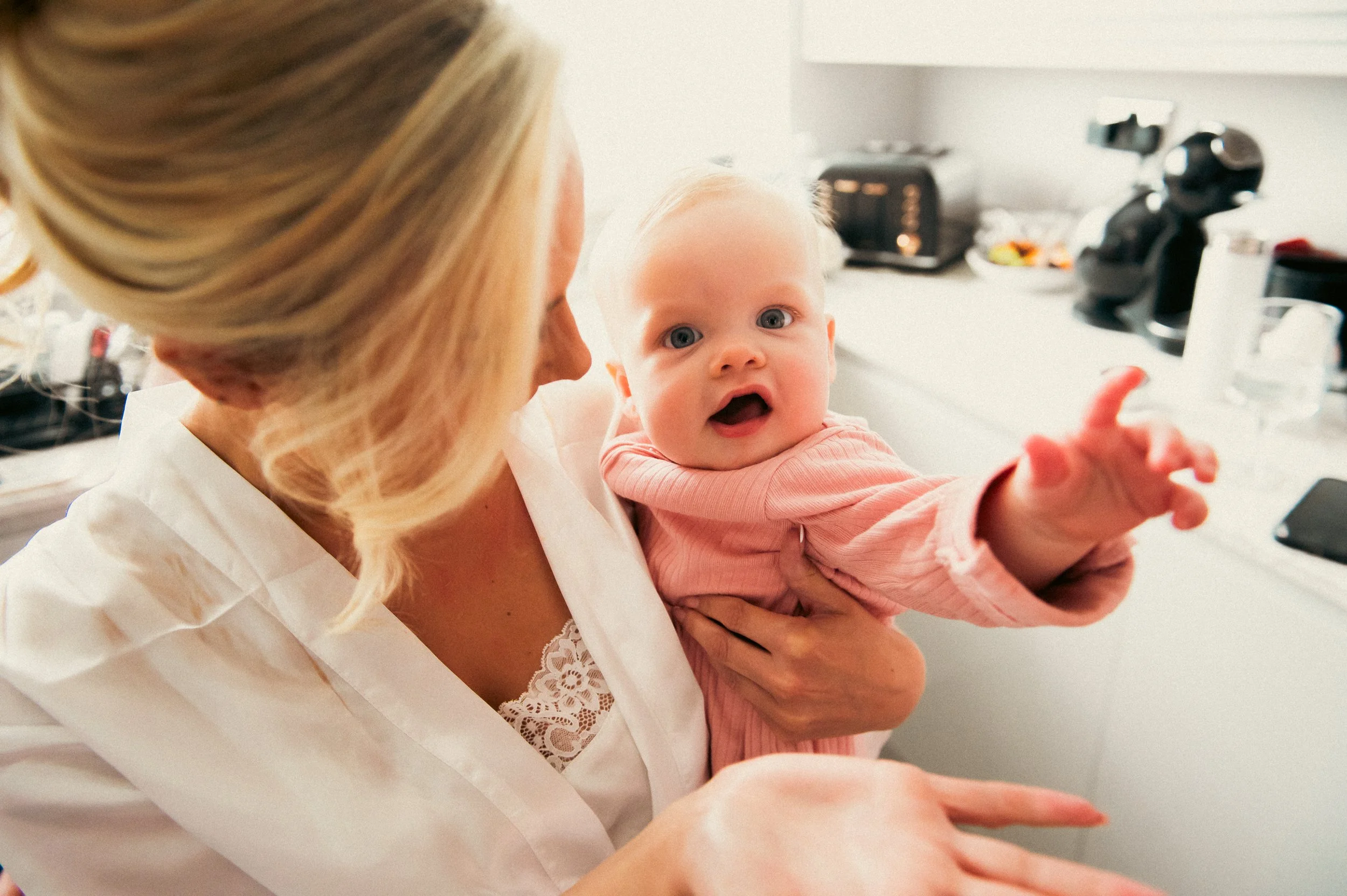 A woman holding a baby in a kitchen, with the baby reaching out towards the camera, looking surprised or curious.