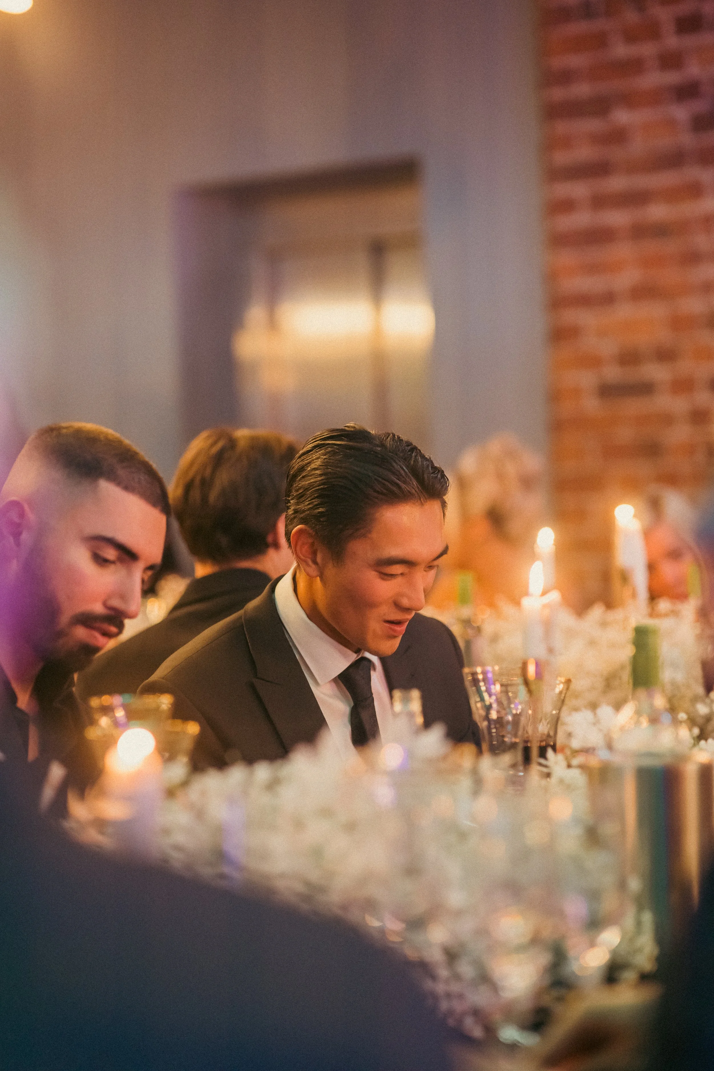 People seated at a formal dining table with candles and floral centerpieces, dressed in formal attire, in an indoor setting with brick and wall decor.