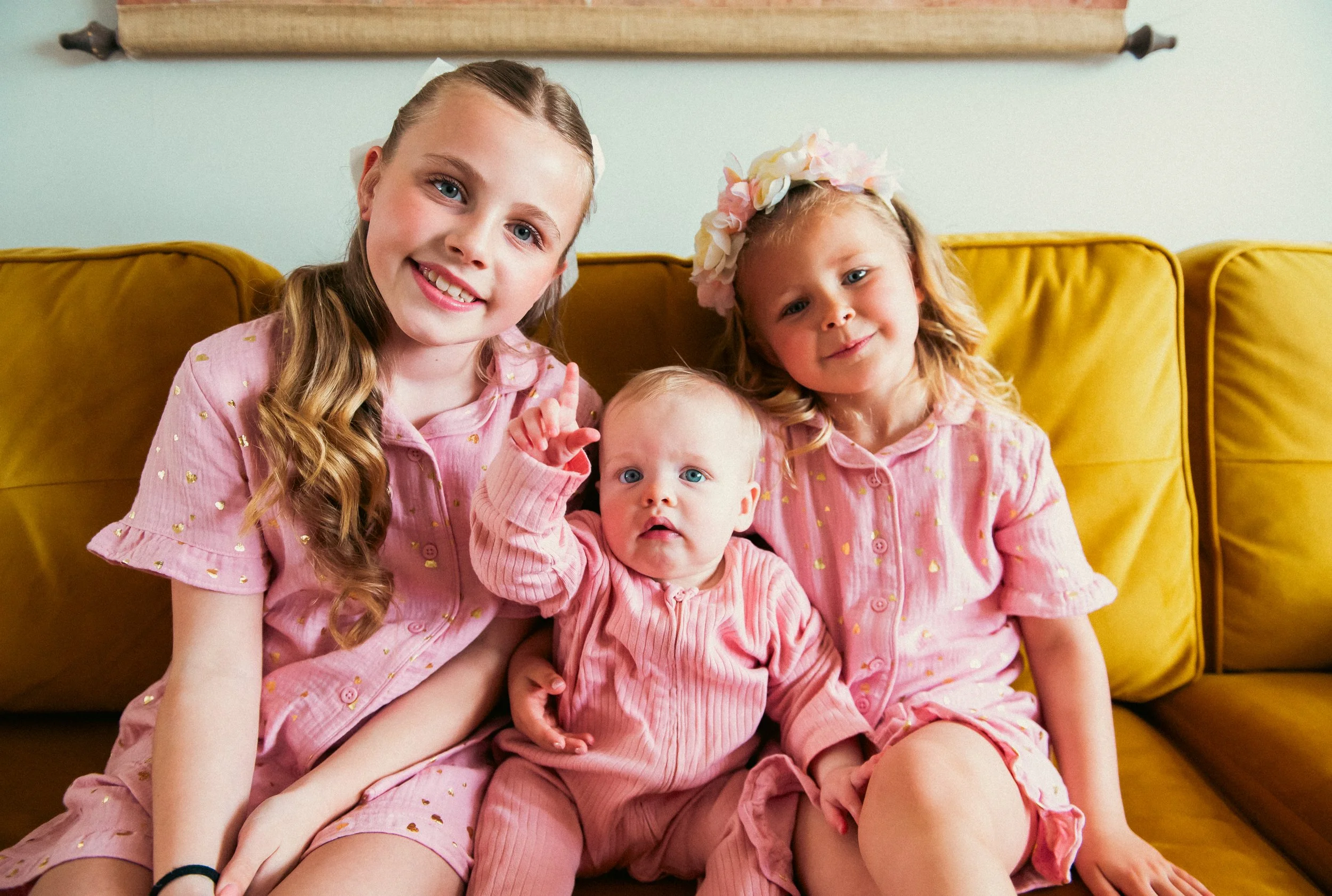 Three young girls sitting on a yellow couch, smiling at the camera. The girl on the left has long, wavy brown hair and is wearing a pink dress with gold accents. The girl in the middle is a baby with blonde hair, wearing a pink outfit, making a hand 