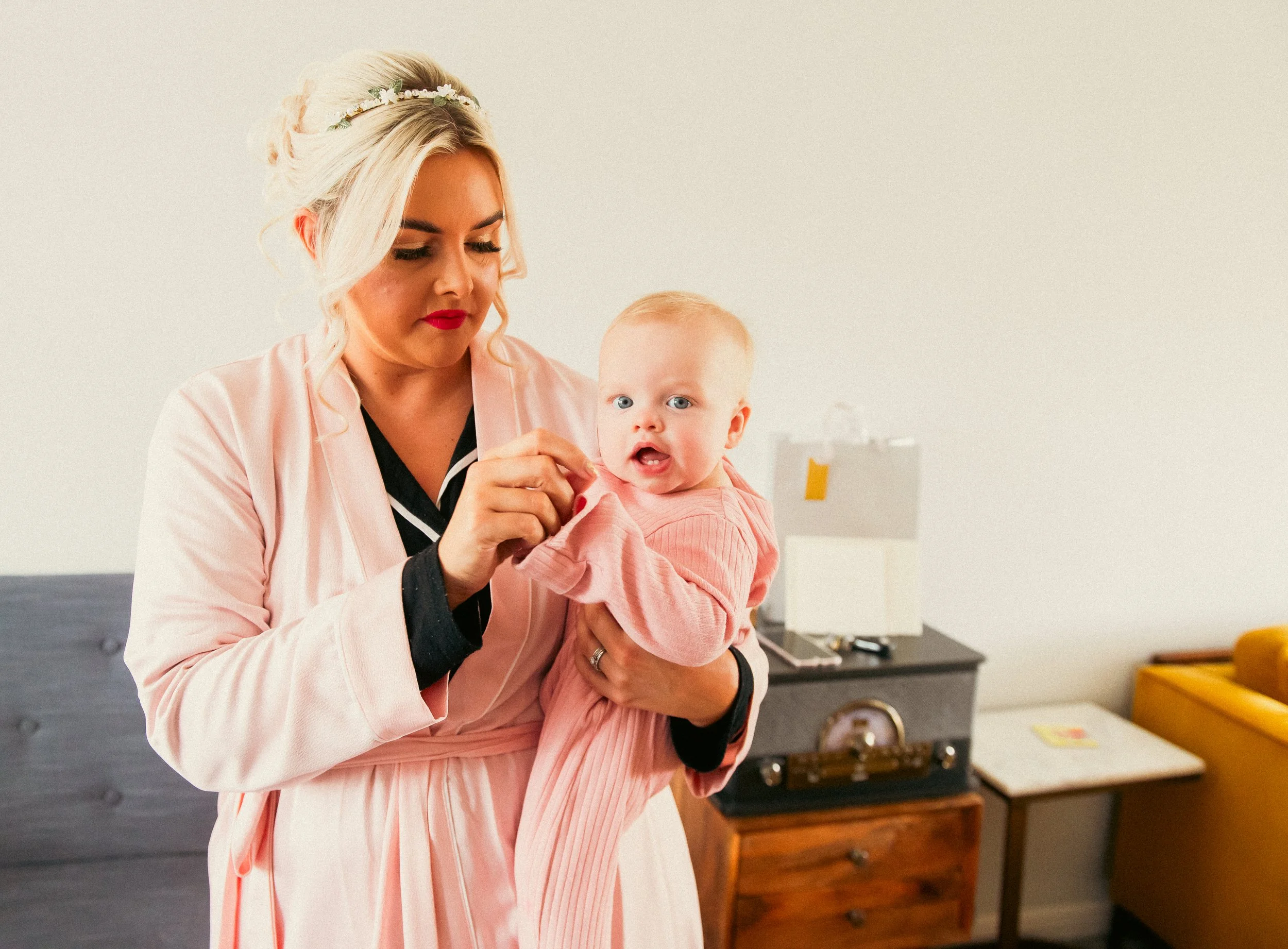A woman with blonde hair in an updo, wearing a pink robe and red lipstick, holding a young girl with blonde hair and blue eyes, dressed in pink. The woman is adjusting the girl's sleeve.