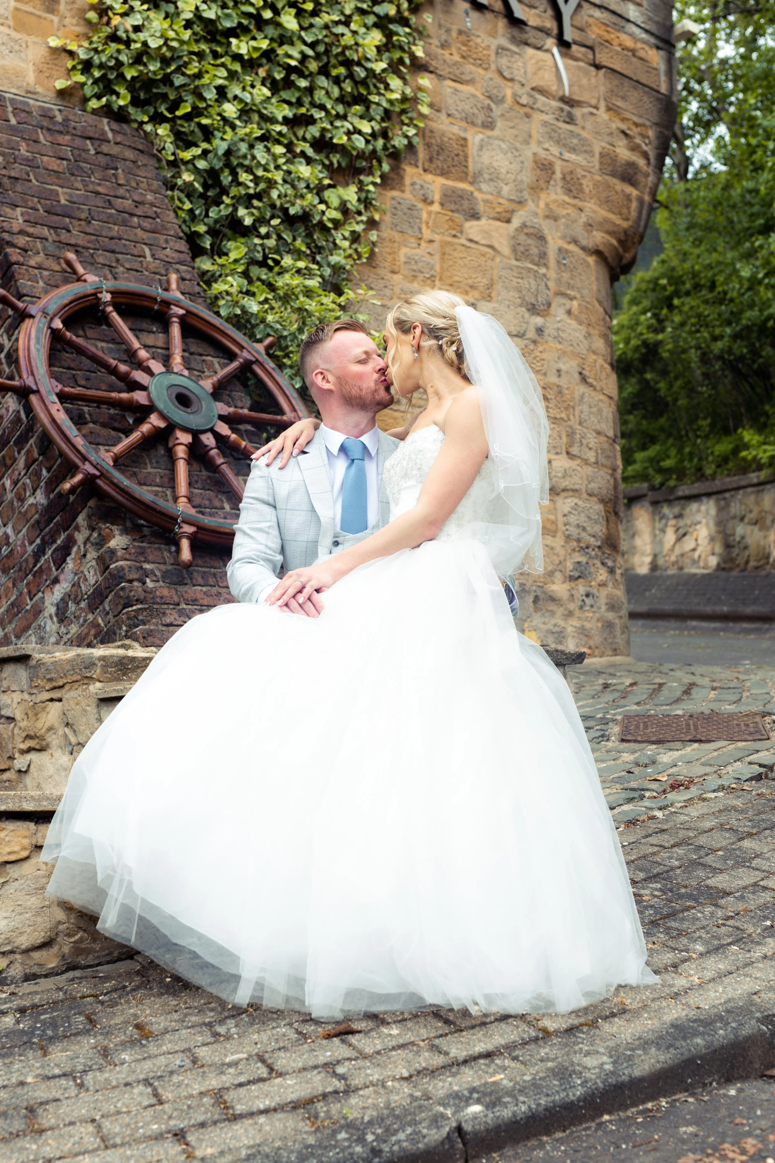 A bride and groom sharing a kiss on a cobblestone street in front of a brick wall with a ship's wheel decoration, surrounded by greenery.