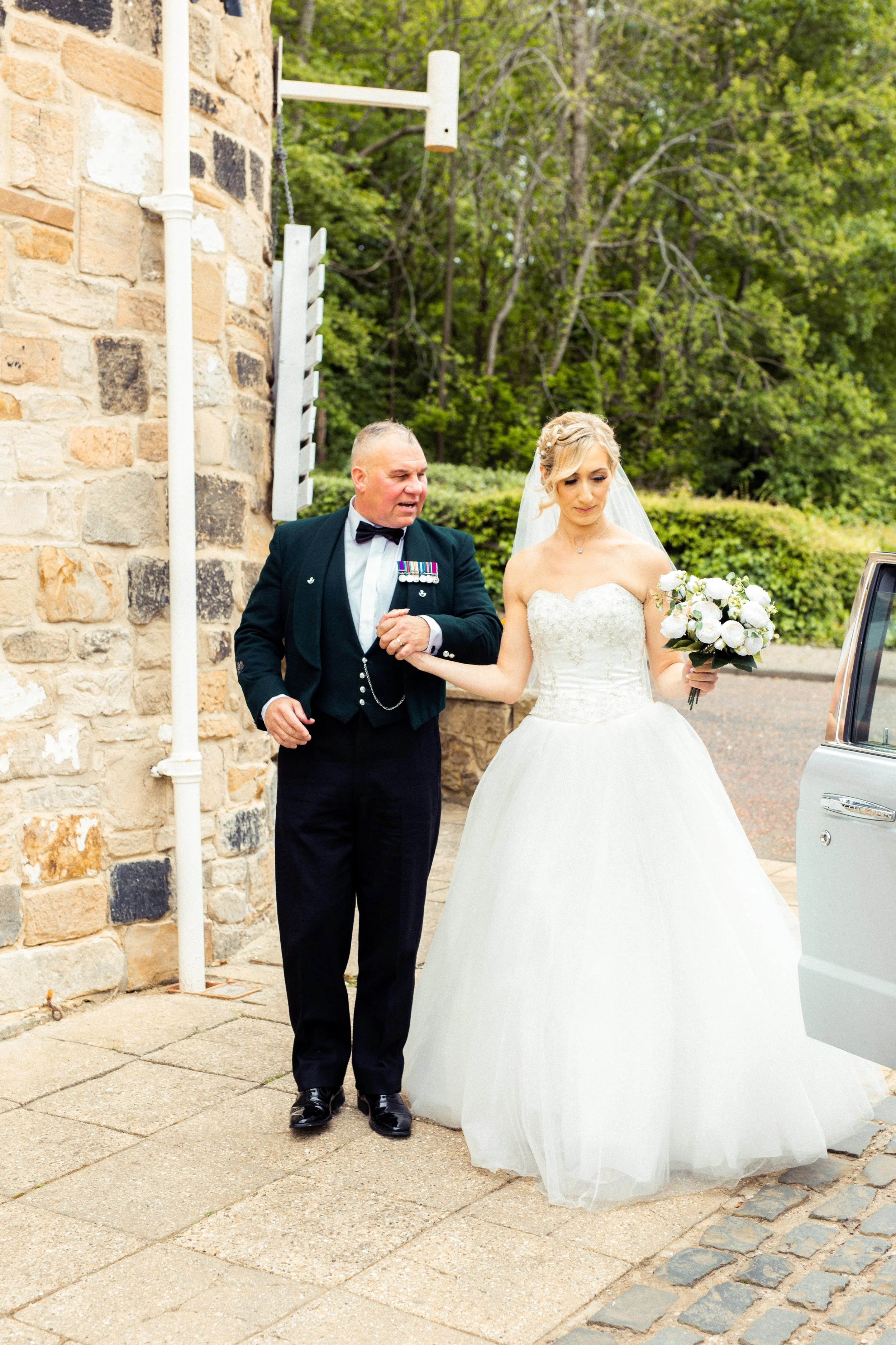 A bride with a bouquet of white flowers being assisted by an older man in a military uniform outside a stone building with green trees in the background.