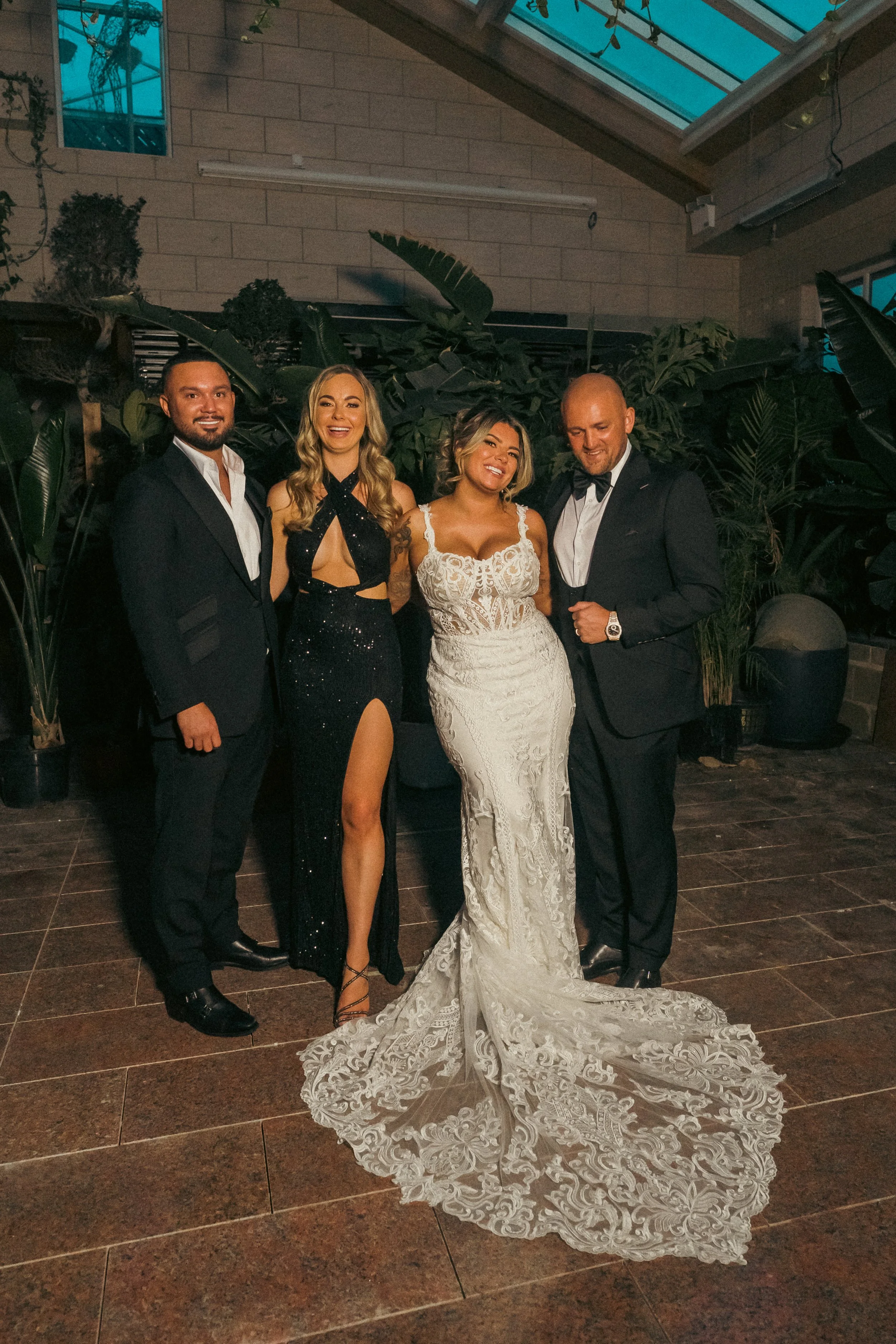 Four people dressed in formal attire at an indoor event with plants in the background. The woman in the center wears a white lace wedding dress with a long train, and the others wear black tuxedos and a black evening gown.