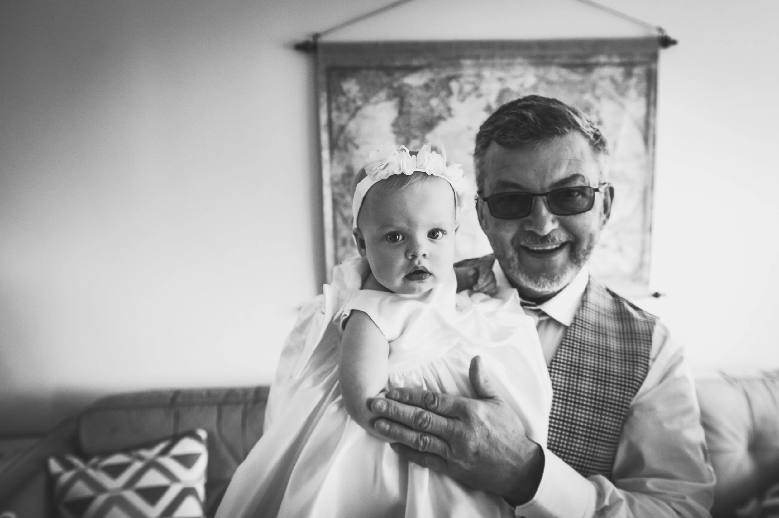 An older man with glasses holding a young girl in a dress and bow on her head, smiling indoors.