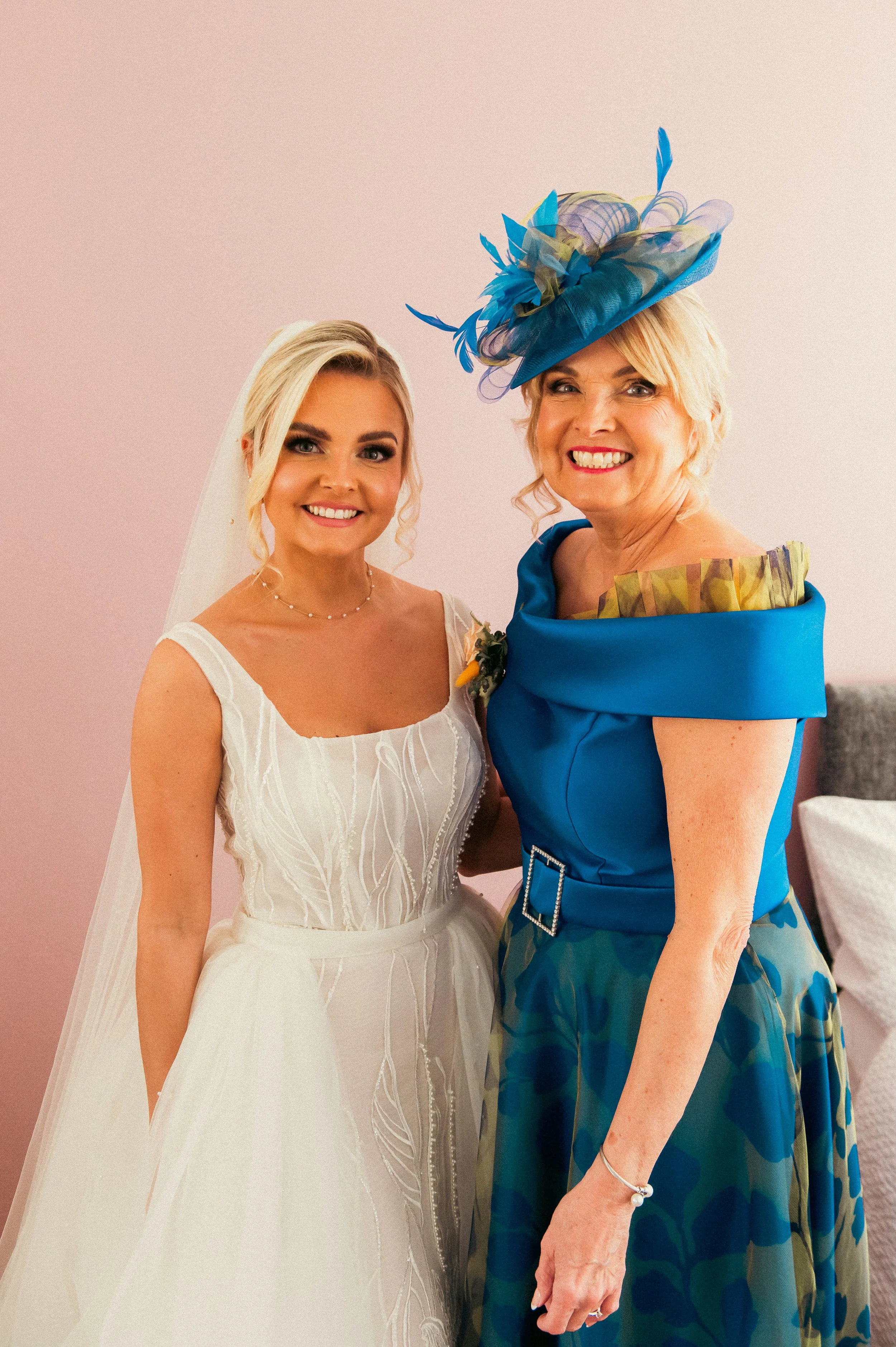 A bride in a white wedding dress posing with an older woman wearing a vibrant blue dress and a matching blue hat with decorative feathers, both smiling indoors against a light pink wall.