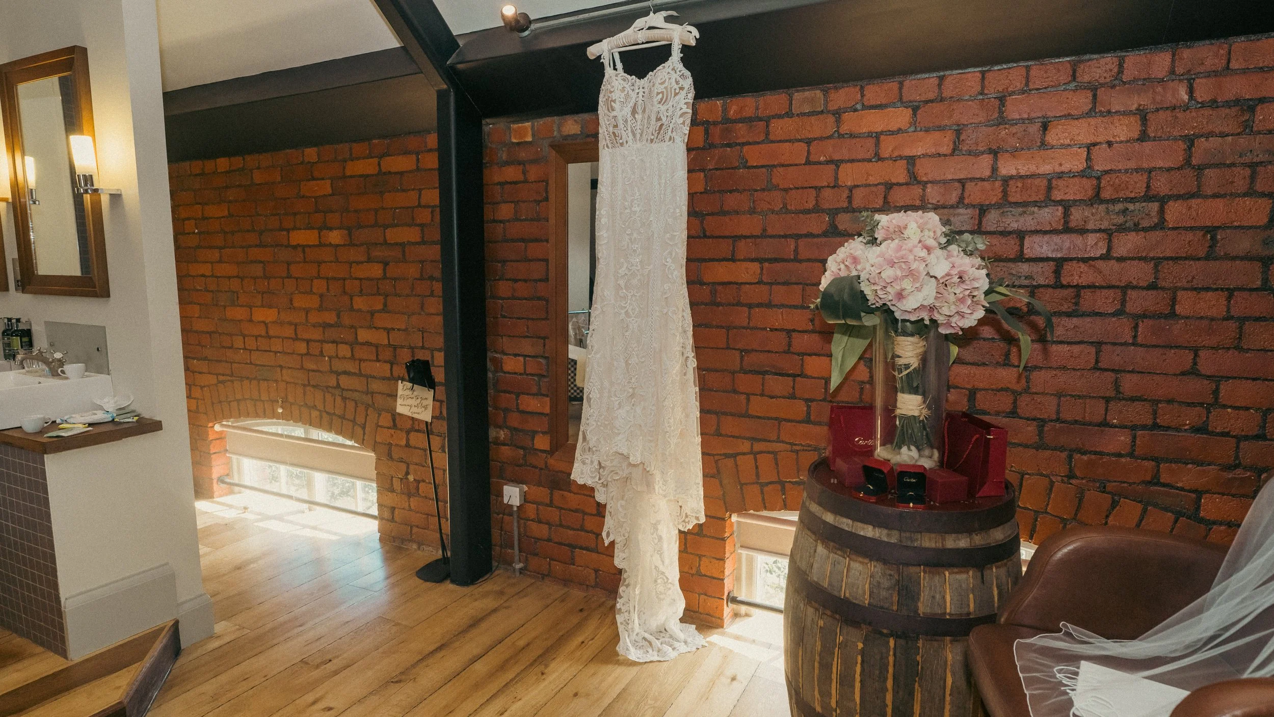 A white lace wedding dress hanging from a black metal frame over a wooden barrel with a large bouquet of pink and white flowers and red gift boxes on top. The setting features a brick wall background and hardwood floors.
