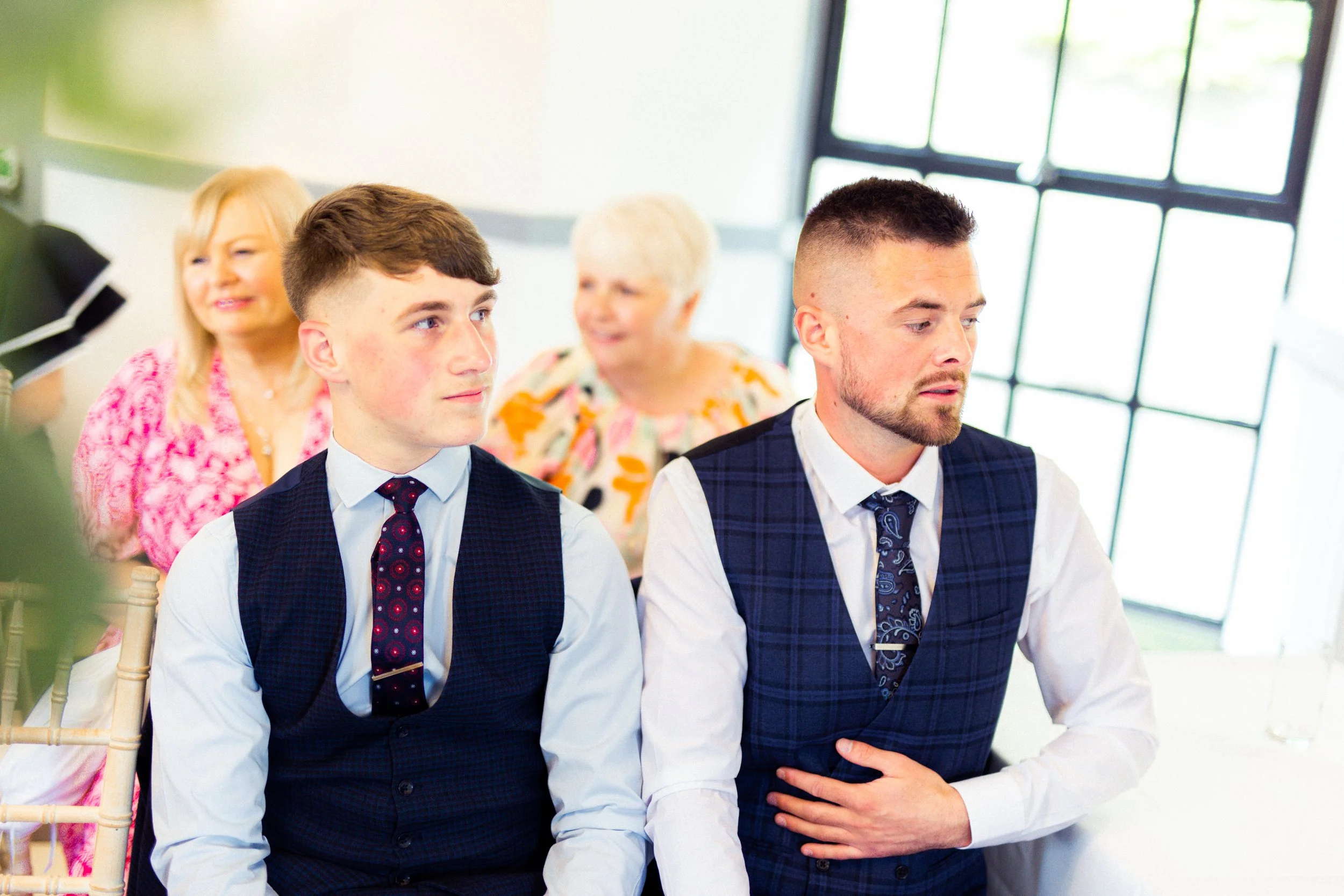 People attending a formal event, including two young men in suits and three older women, sitting at a table near large windows.