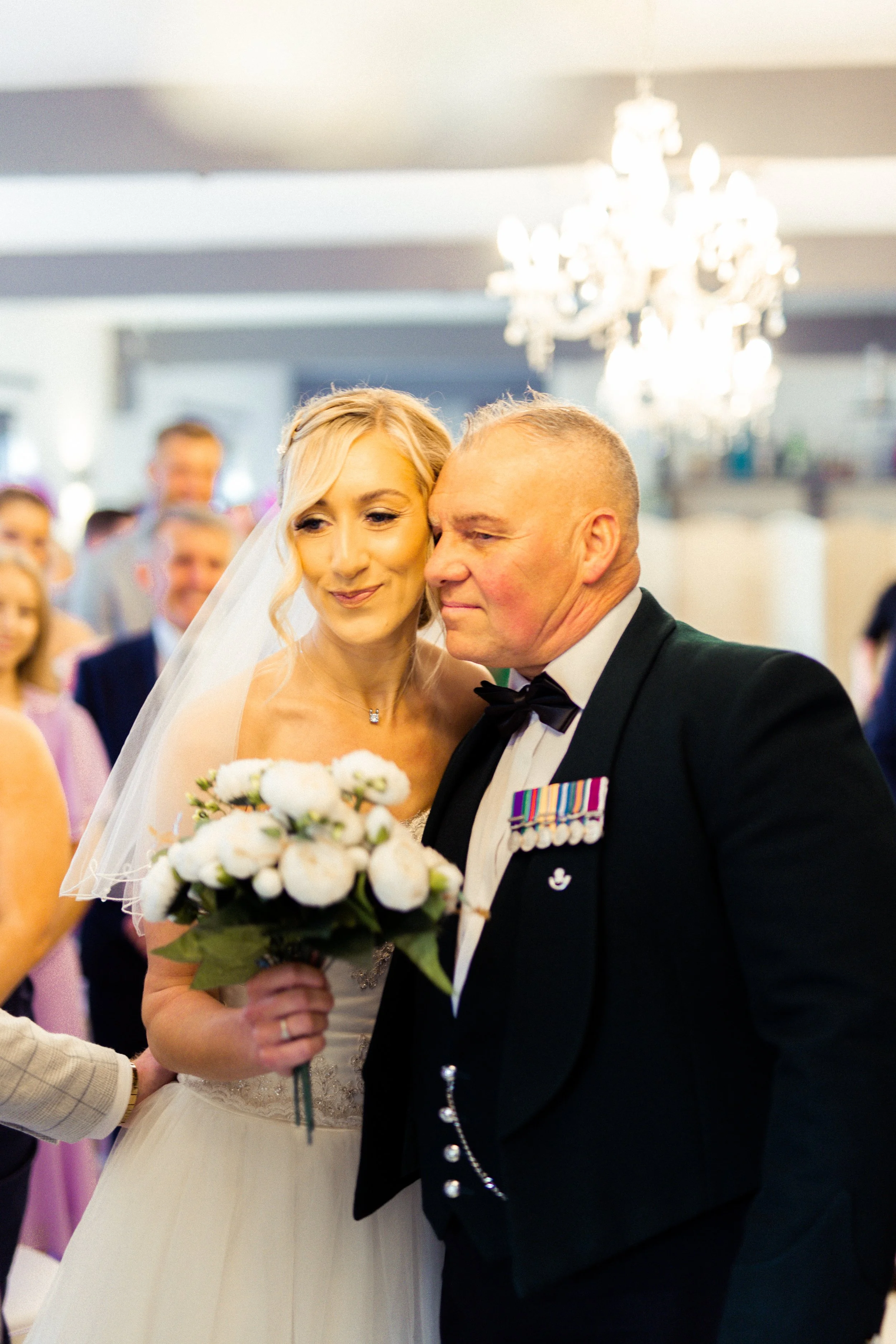 Bride and groom sharing a moment during their wedding, with the bride holding a bouquet of white flowers, surrounded by wedding guests in a decorated indoor venue.