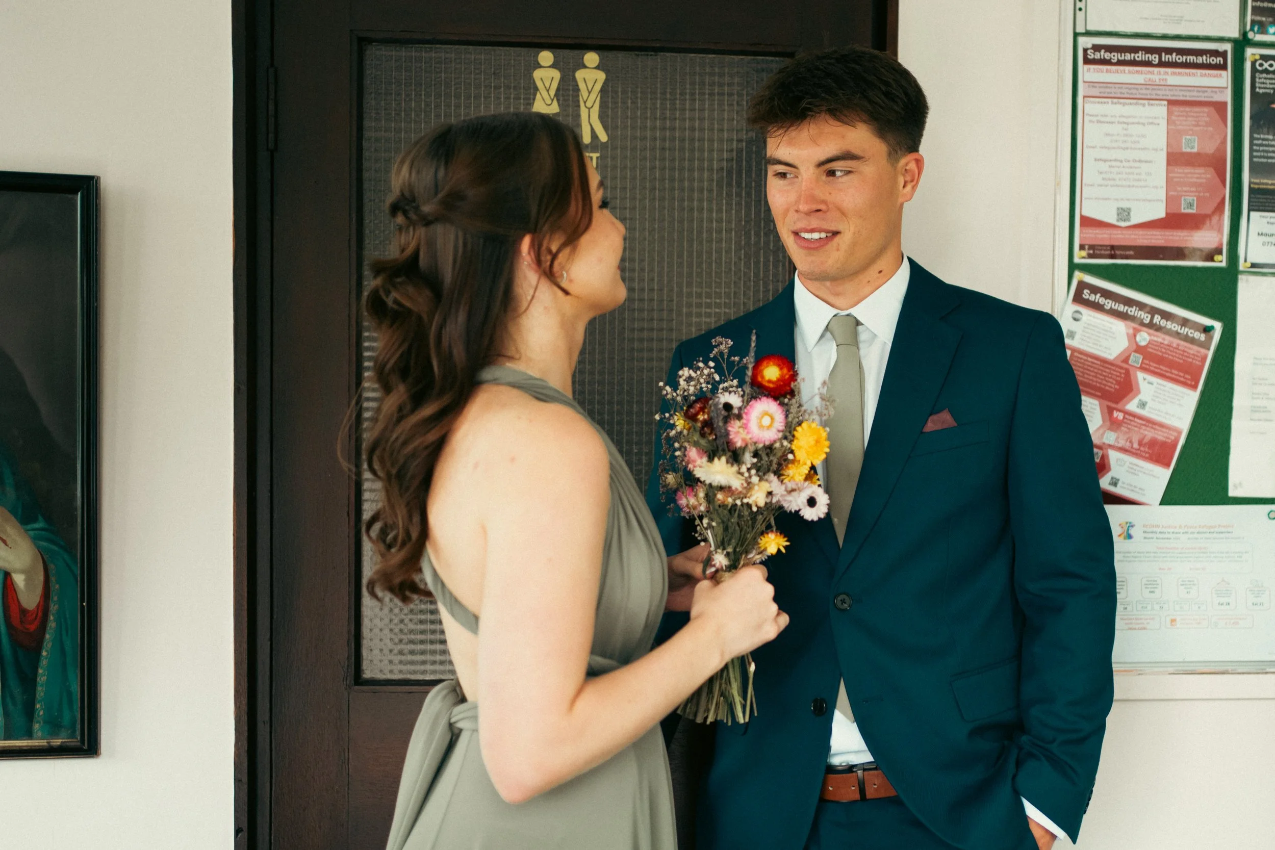 A young woman in a gray dress holding a colorful bouquet of flowers is smiling and looking at a young man in a navy suit with a gray tie, who is smiling back. They are standing indoors near a door with a restroom sign and a bulletin board.