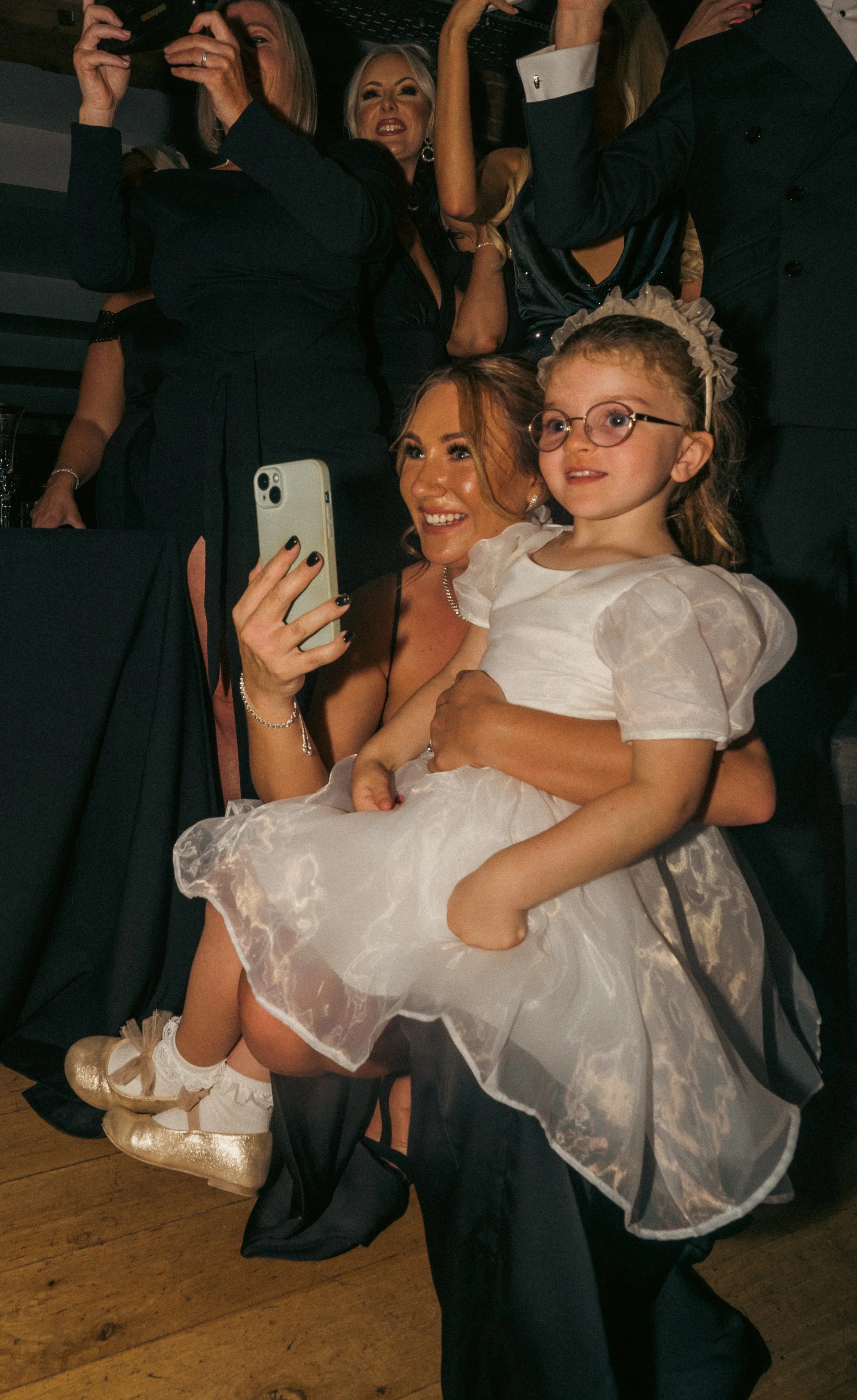 A woman with red hair and a young girl with glasses and a white dress taking a selfie at a celebration or party, surrounded by other people smiling and posing for photos.