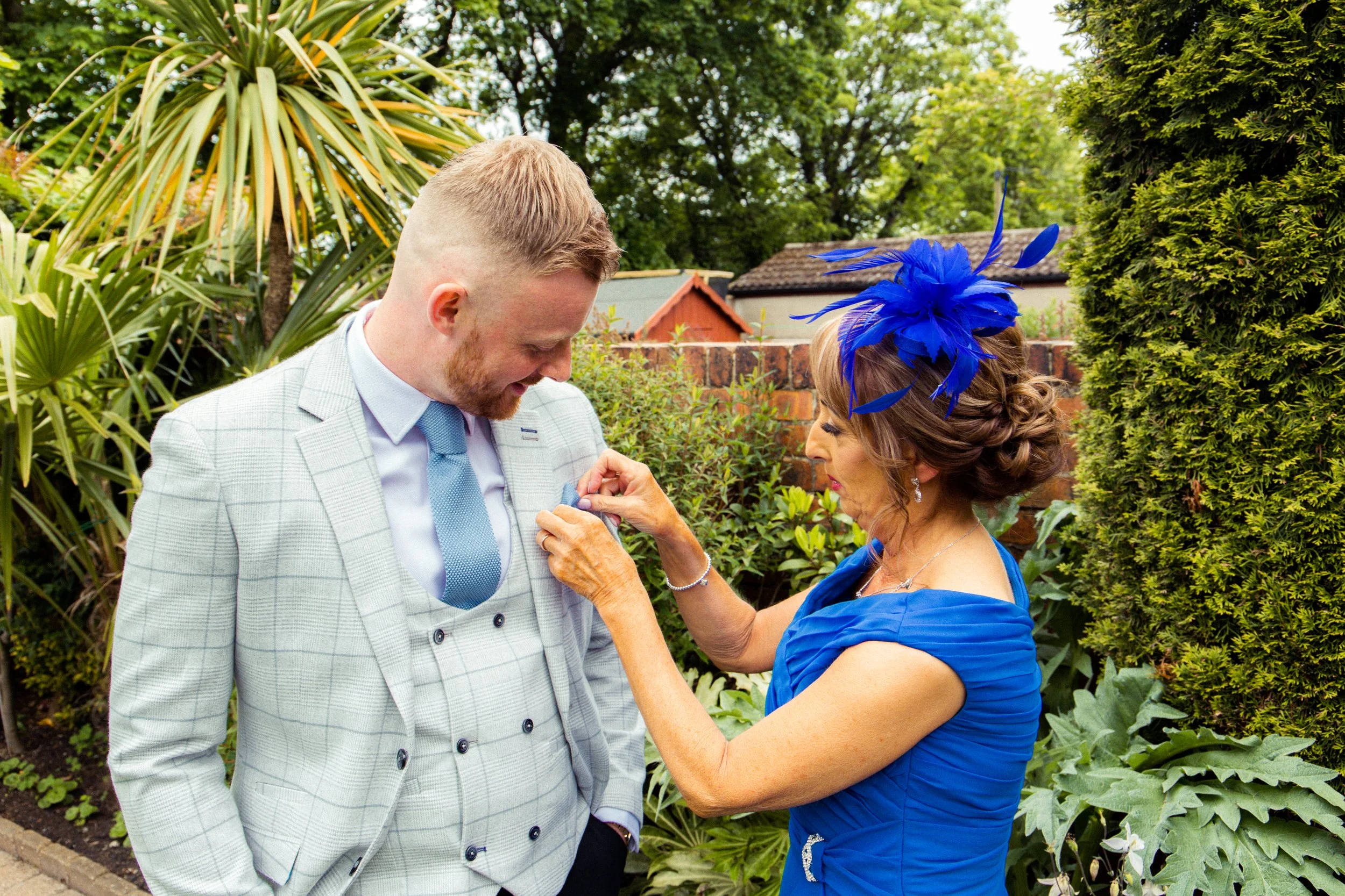 A woman in a blue dress with a blue feathered headpiece pinning a boutonniere on a man in a light-colored suit and tie in a garden.