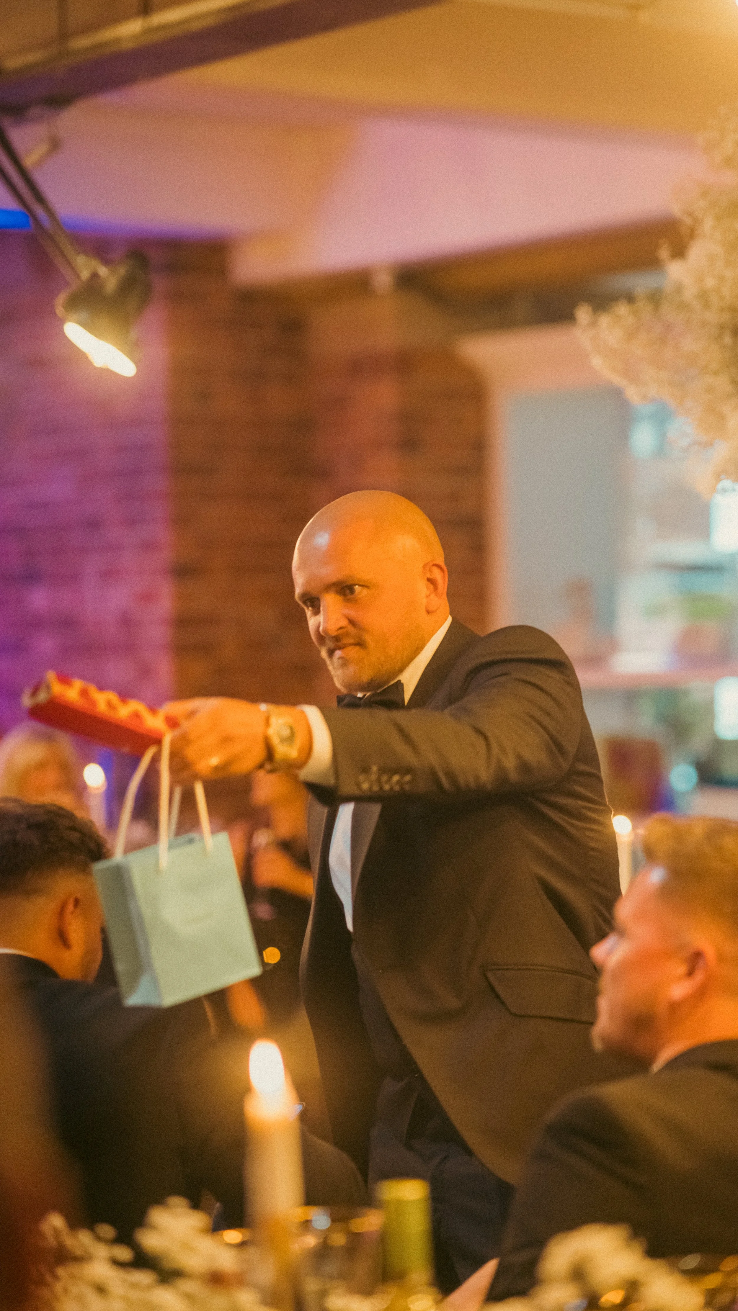 A man in a tuxedo is holding a gift and a pizza box at a formal gathering, with others seated at tables and candles on the table.