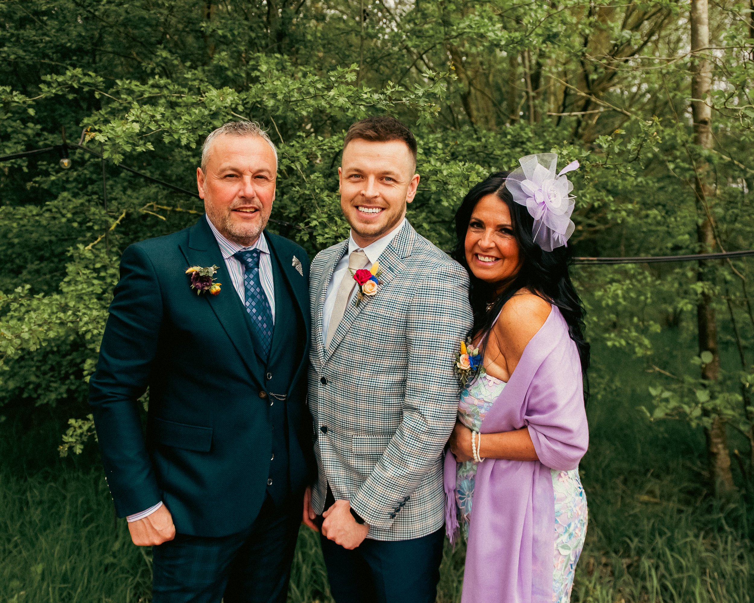 Three people standing outdoors in front of green trees, dressed in formal and festive attire, smiling at the camera.