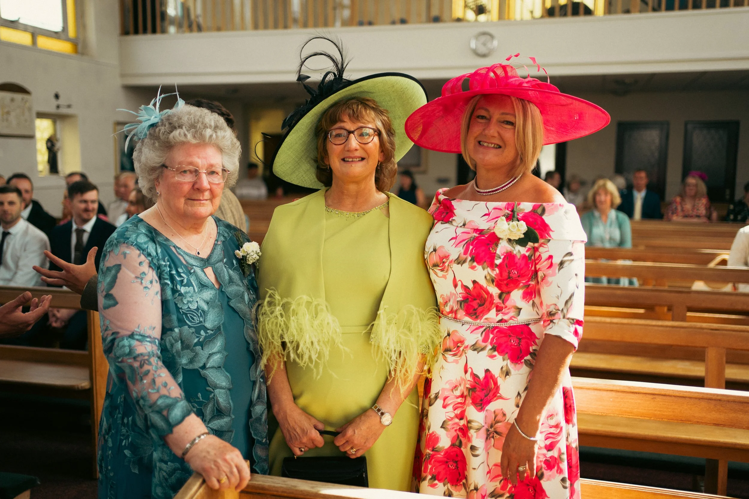 Three women in colorful dresses and large hats standing inside a church during a wedding, with guests seated in pews in the background.
