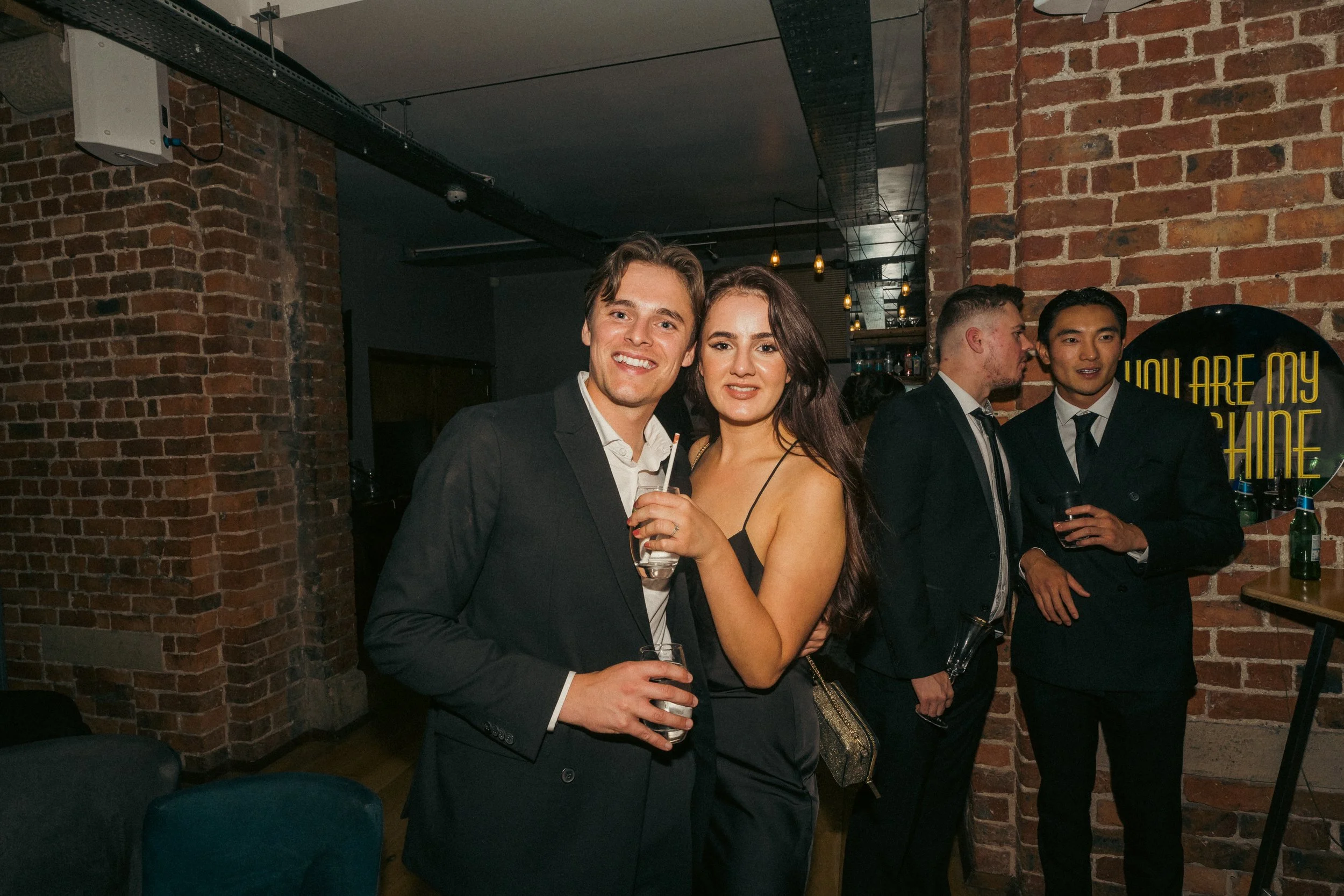 A group of four people at a bar or nightclub, dressed in formal attire, standing near a brick wall, with two women and two men, some holding drinks.