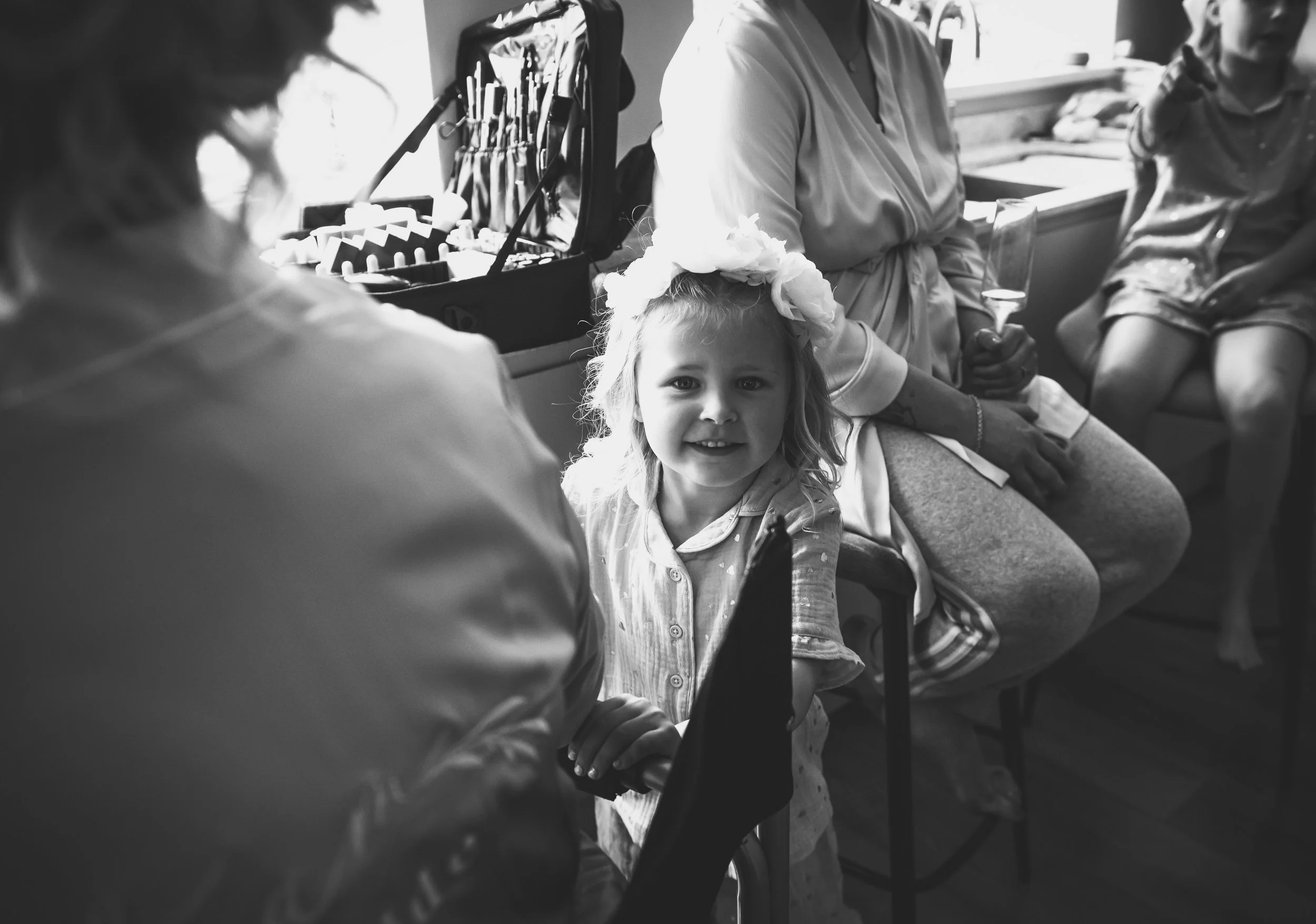 A young girl with a flower crown smiling at the camera, sitting at a social gathering with adults and children, with gift boxes and a drink glass nearby.