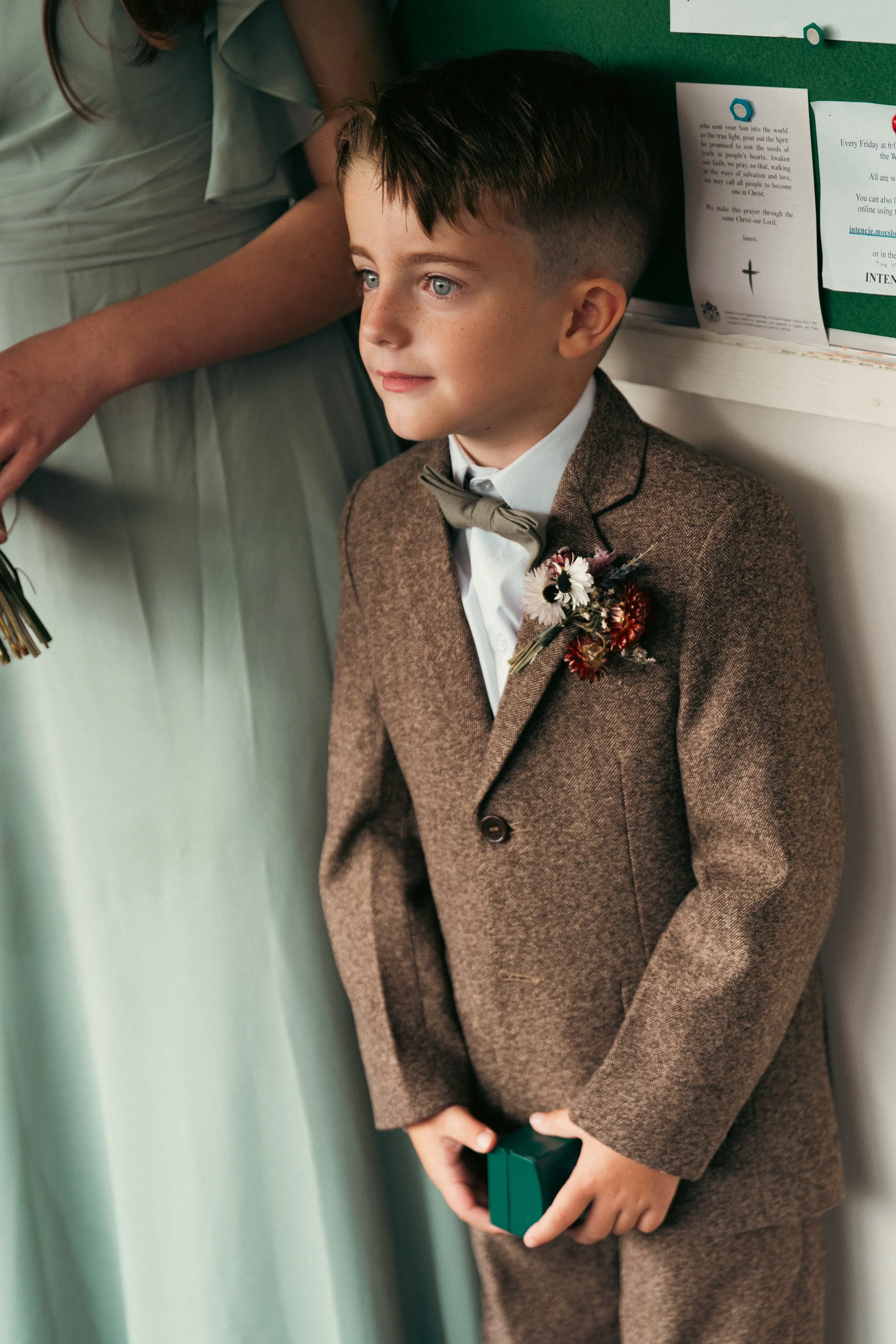 A young boy dressed in a brown suit with a boutonniere, white shirt, and bow tie, holding a small green box, standing indoors near a green bulletin board with papers.
