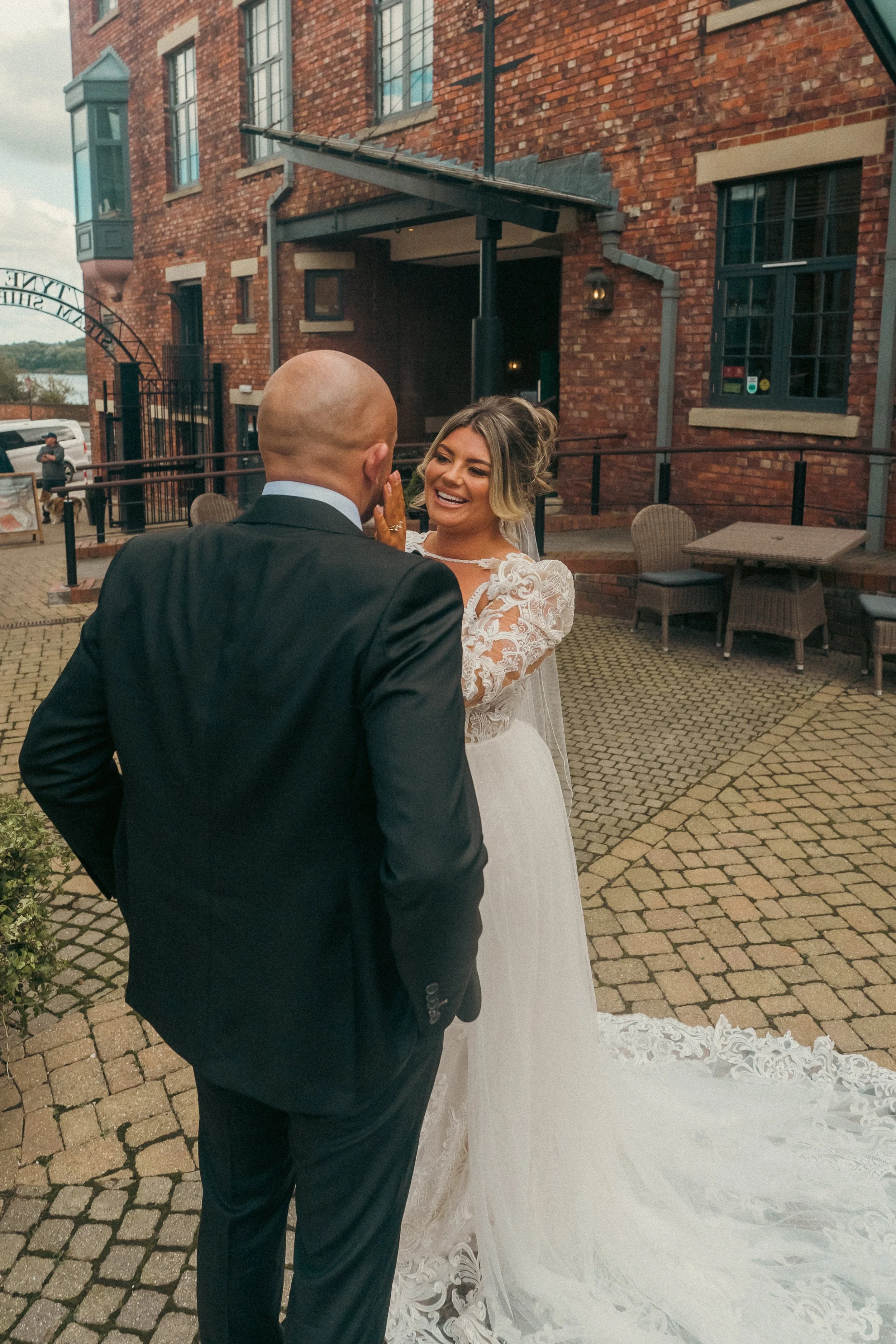 A bride smiling and touching a groom's face outside a brick building on her wedding day.