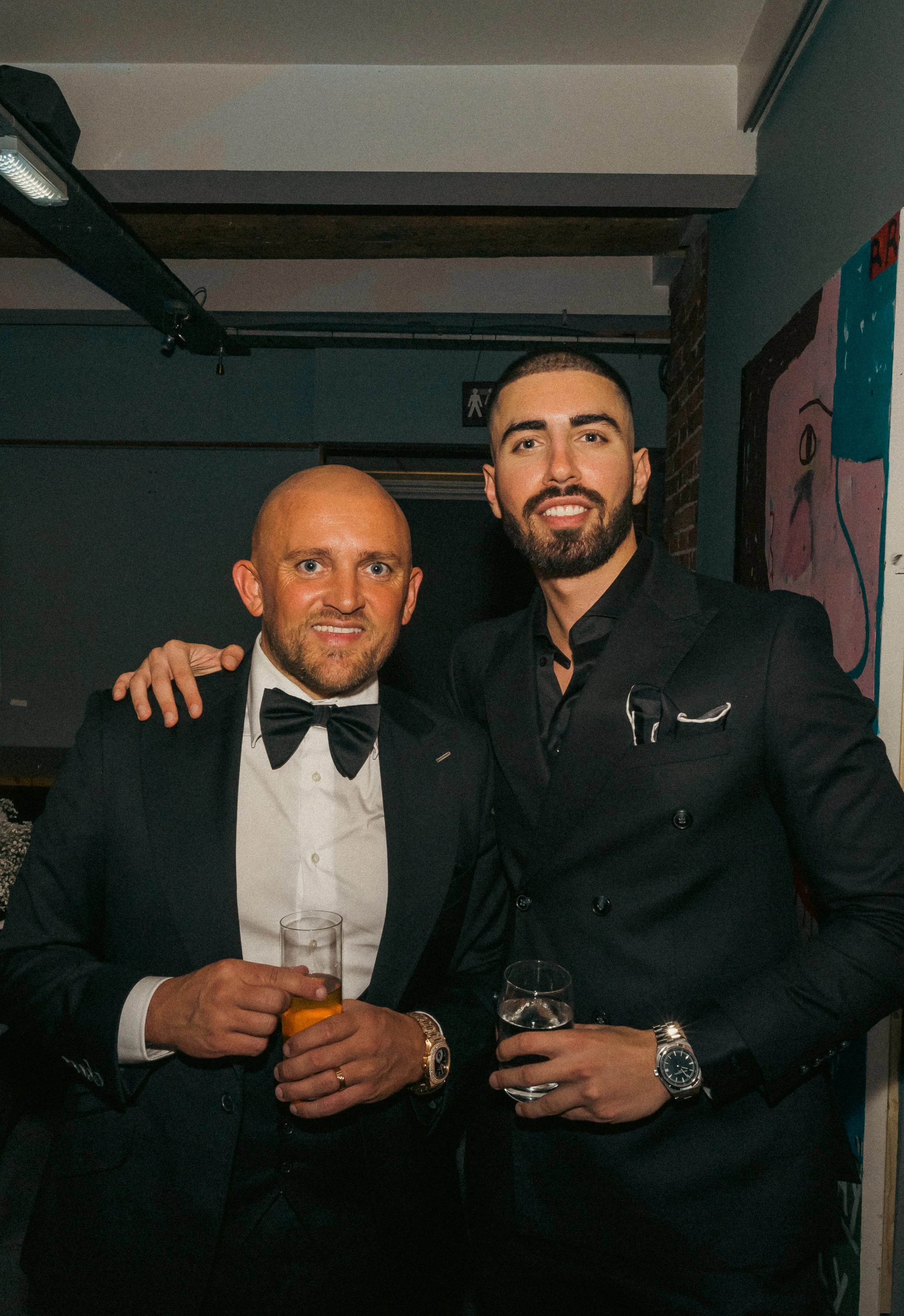 Two men in tuxedos posing together at an indoor event, each holding a drink, smiling at the camera.