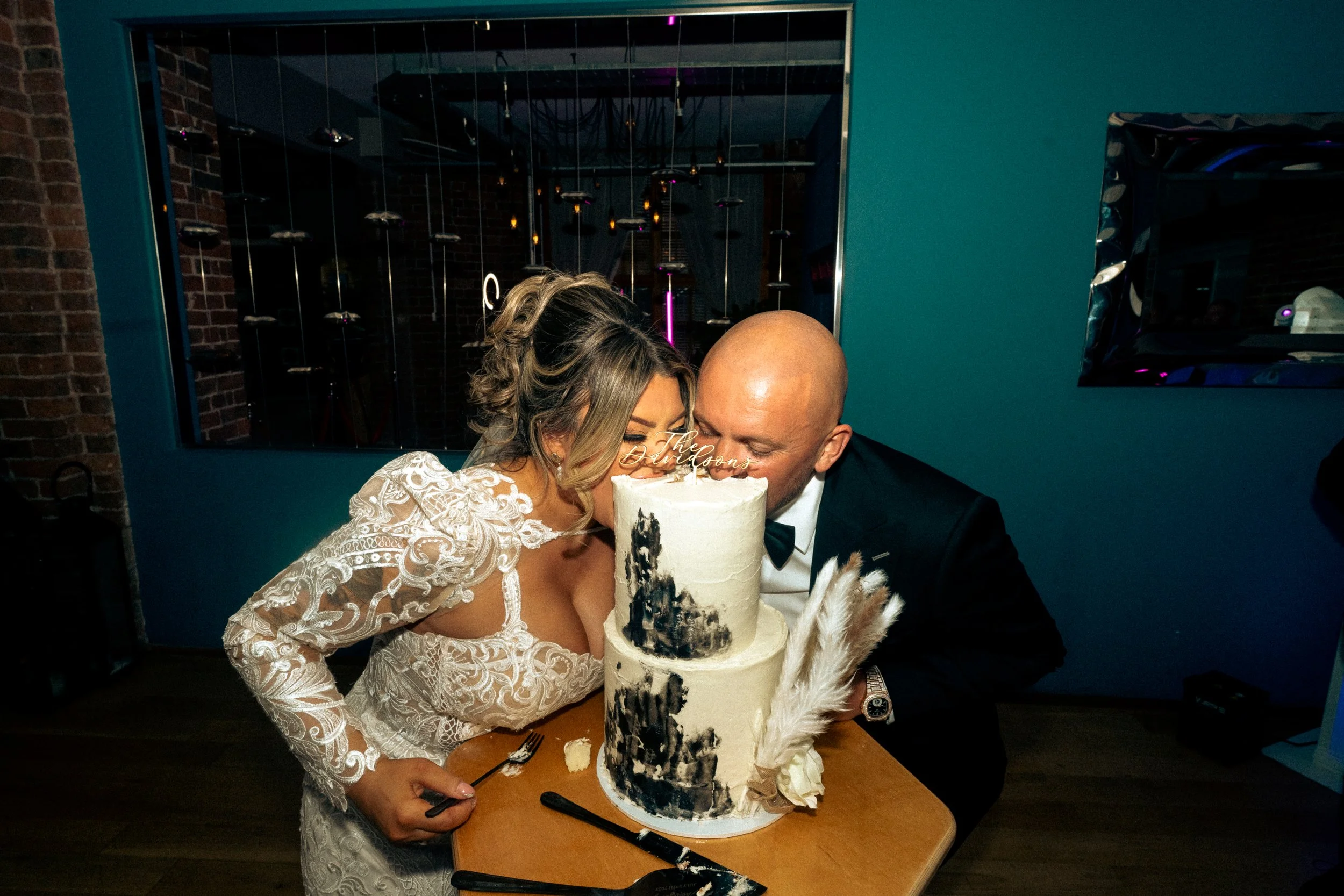 A bride and groom wearing wedding attire, leaning over a wedding cake to kiss, at their wedding celebration.