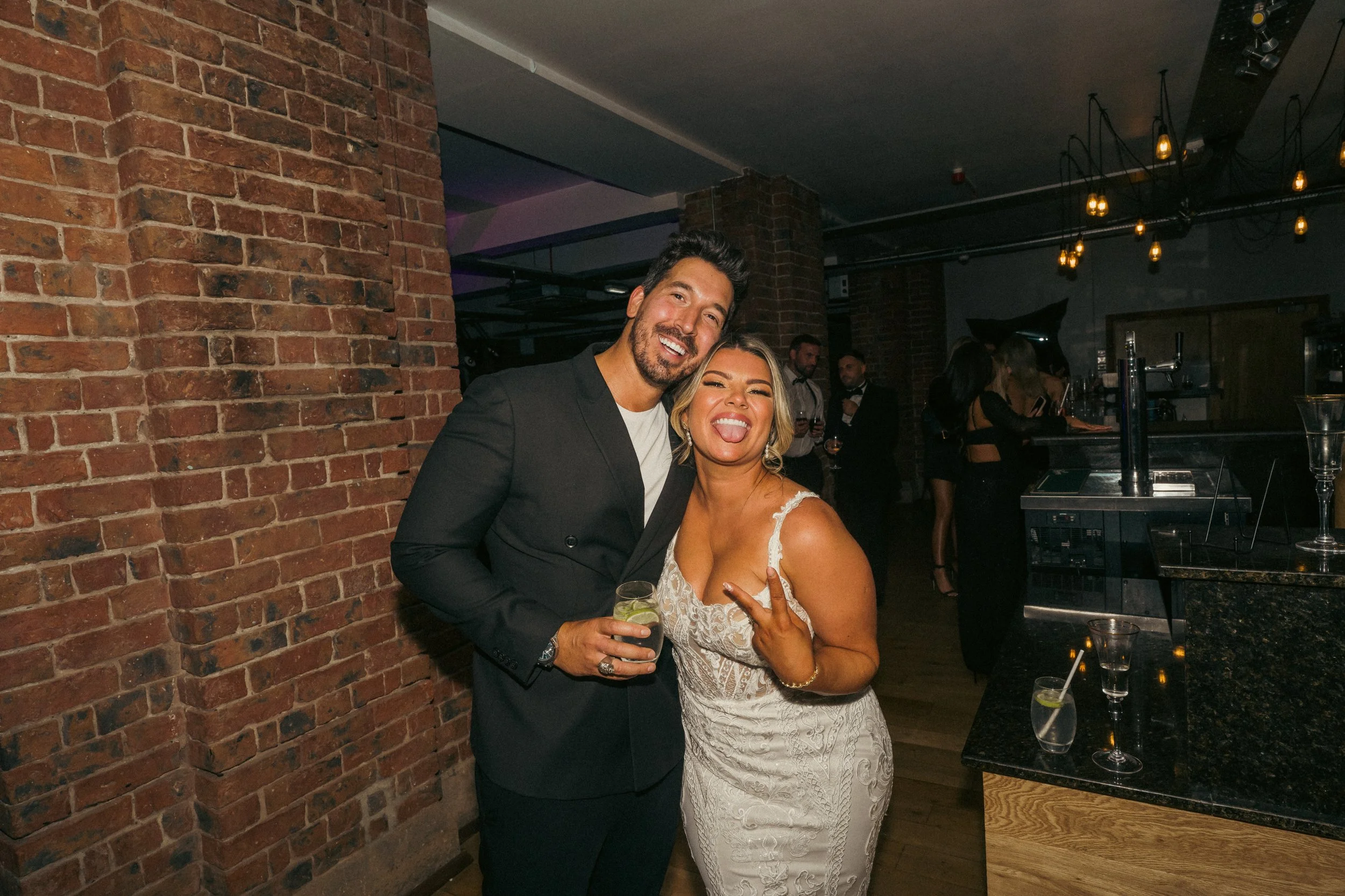 A man and woman smiling and posing together at a social event in a bar or nightclub, with drinks in hand. The man is wearing a black suit, and the woman is in a white lace dress, making a peace sign.