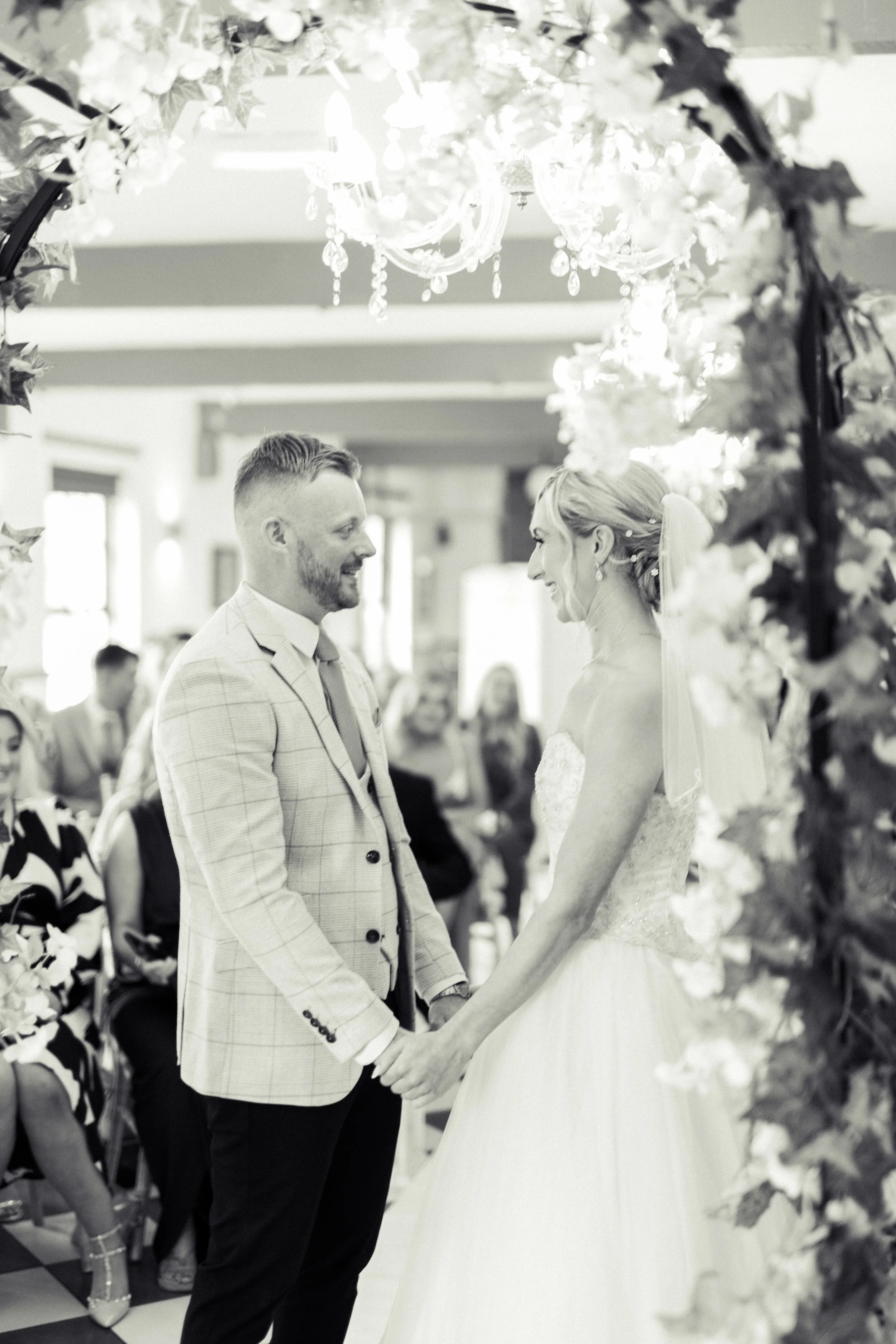 A bride and groom exchanging vows during their wedding ceremony, surrounded by floral decorations and seated guests, in a well-lit indoor venue.