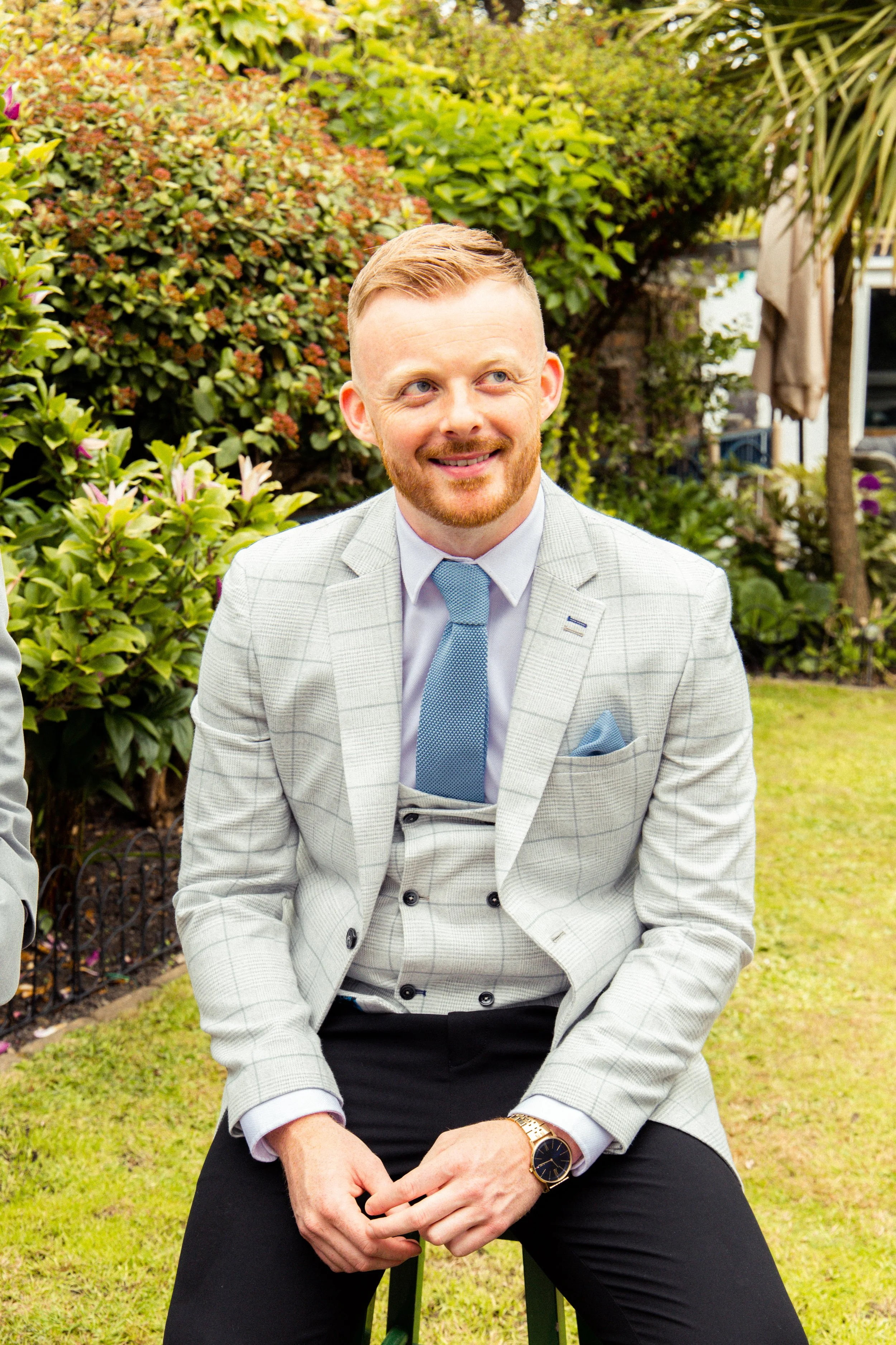 A man with red hair and a beard, dressed in a light gray plaid suit with a blue tie, sitting outdoors on a grassy area surrounded by green bushes and trees, smiling and looking to his left.