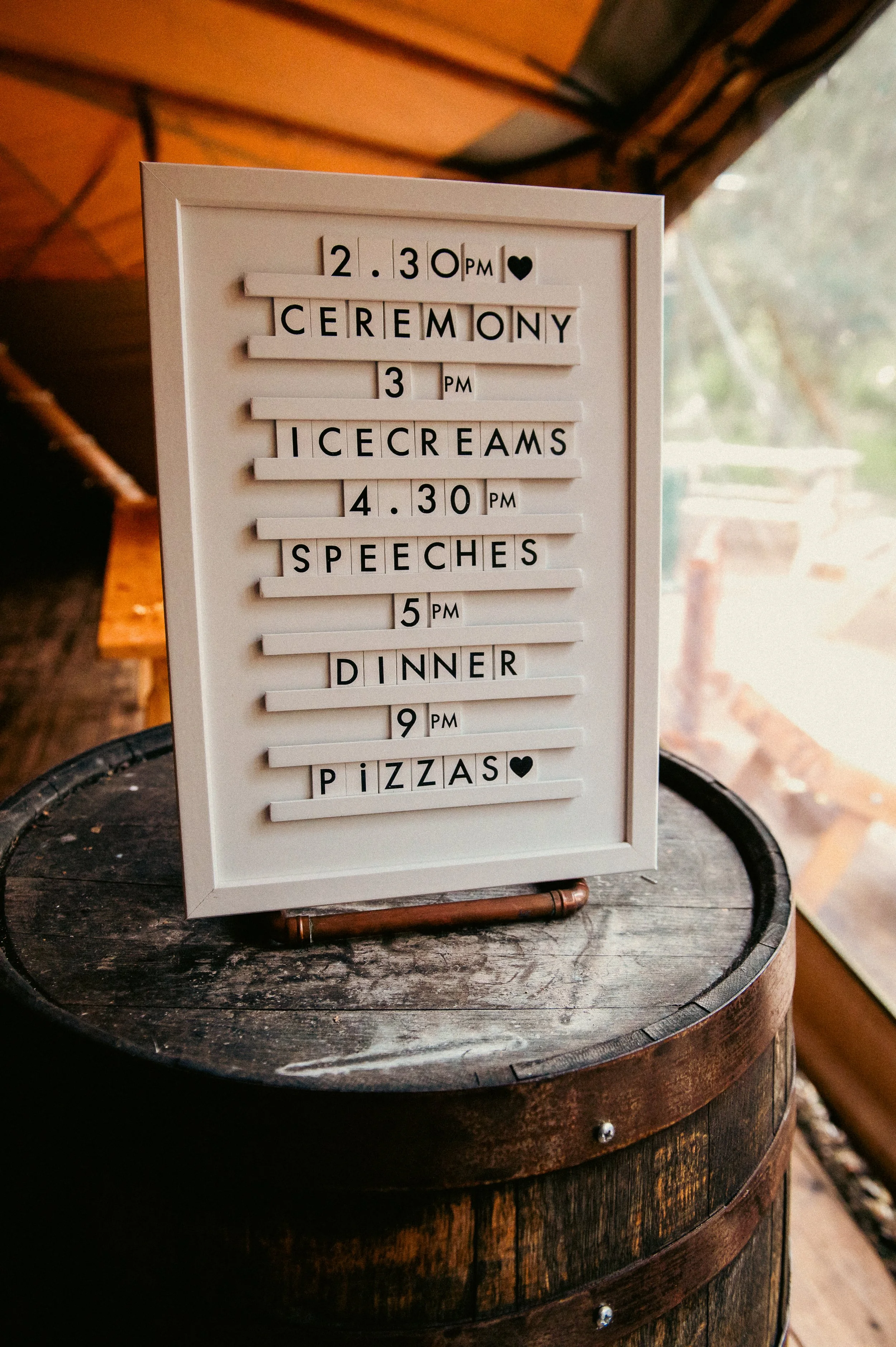 A white letter board with black letters displaying a wedding schedule, including ceremony at 2:30 PM, ice creams at 3 PM, speeches at 4:30 PM, dinner at 5 PM, and pizzas at 9 PM, placed on a wooden barrel inside a rustic tent.