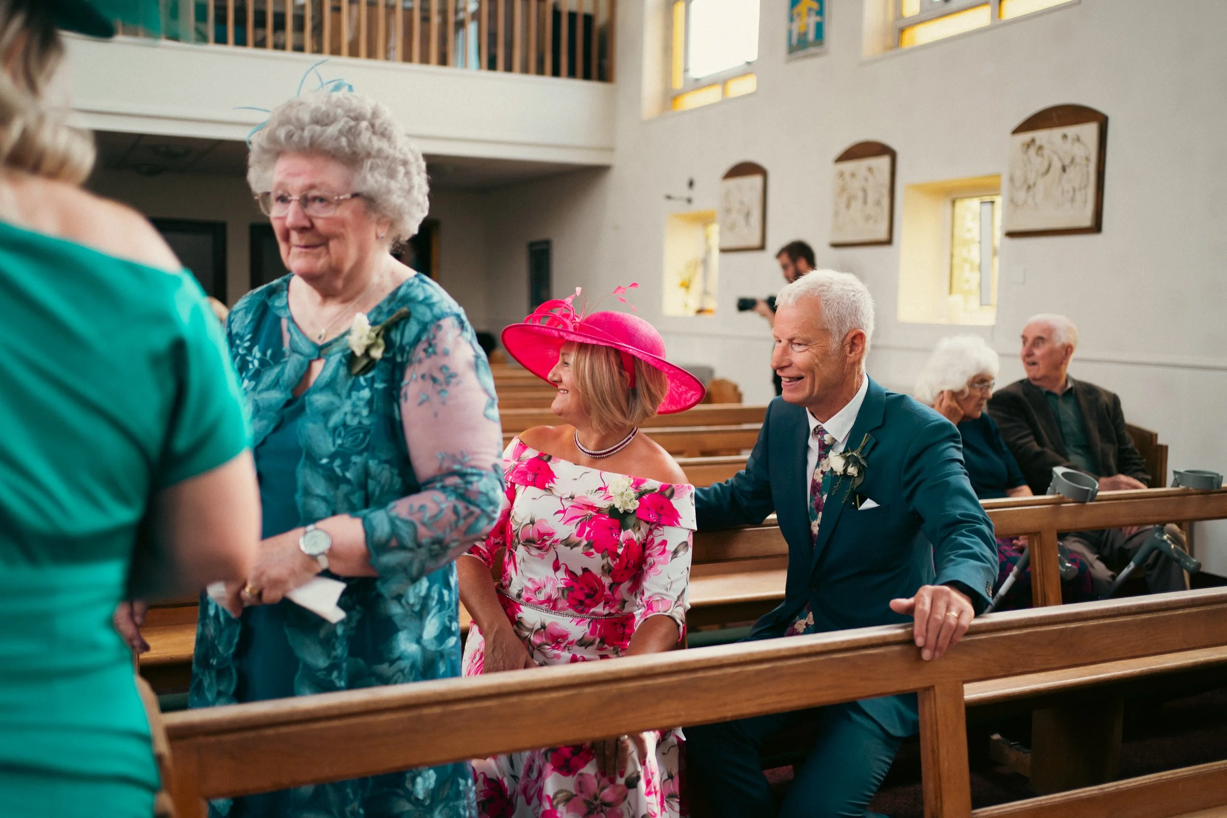 People attending a wedding ceremony inside a church, sitting on wooden pews and engaging with each other. Two elderly women and a man in a suit are visible, along with a woman in a bright floral dress and large pink hat.