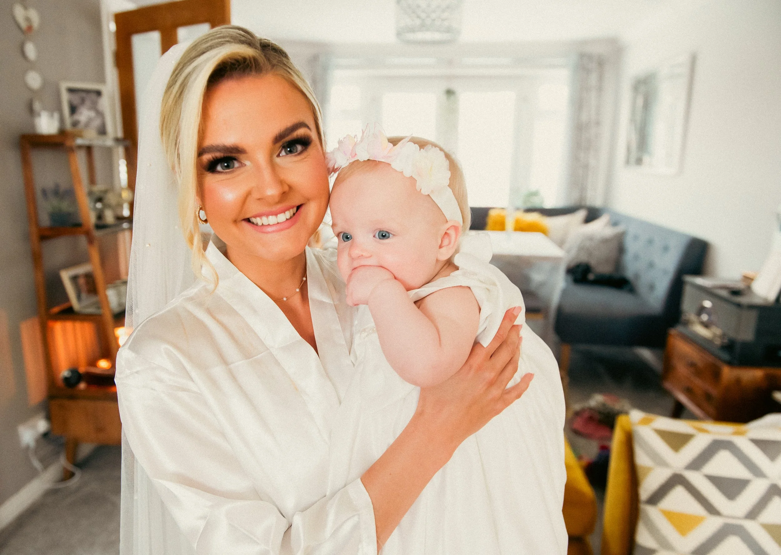 A smiling woman in a white robe holding a baby girl with a flower headband in a bright living room.